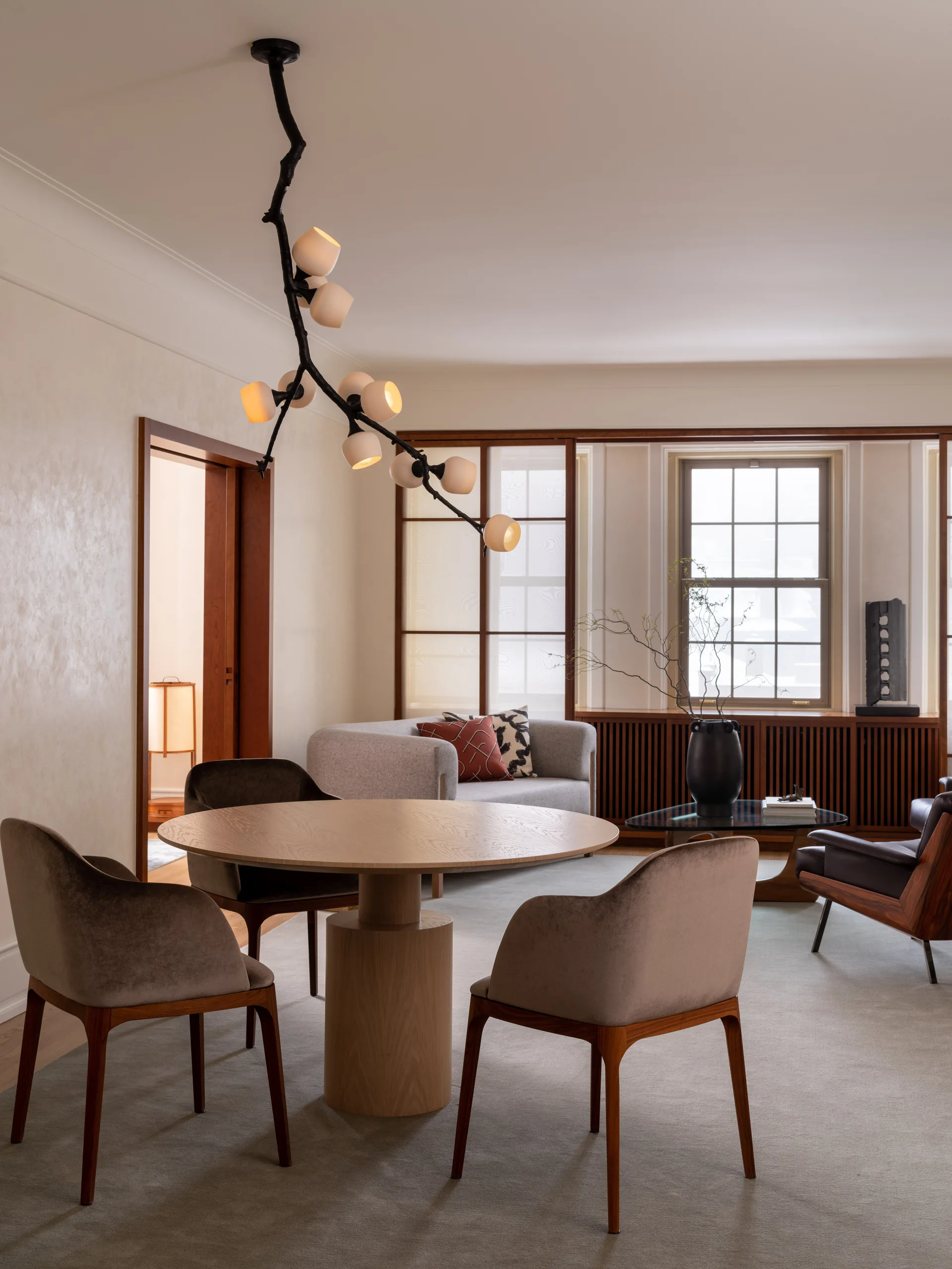 A circular wood dining table is surrounded by grey upholstered armchairs beneath a black branch chandelier in a warm neutral room.
