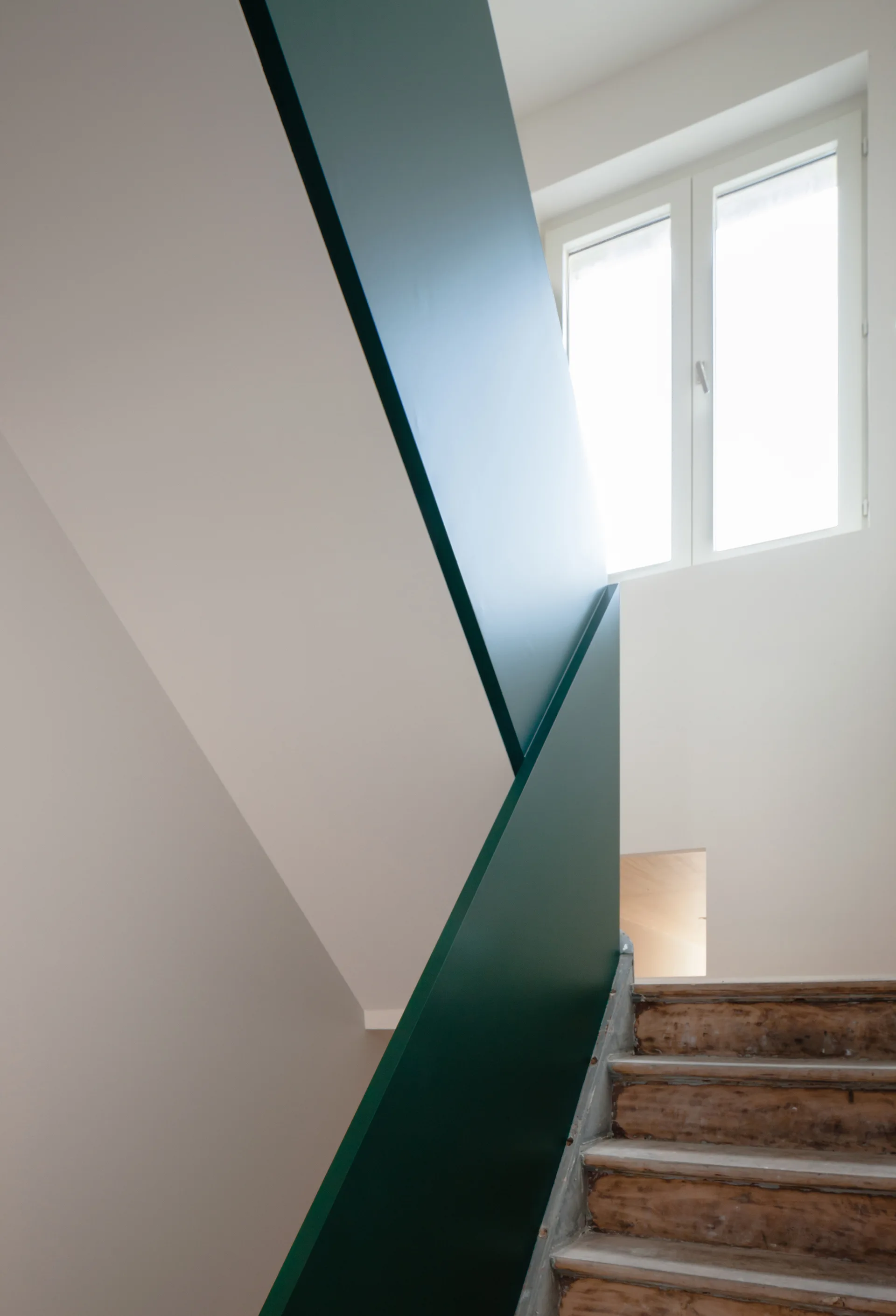 A minimalist interior staircase features unfinished wooden steps alongside a solid deep green painted geometric guardrail beneath a white-framed window.