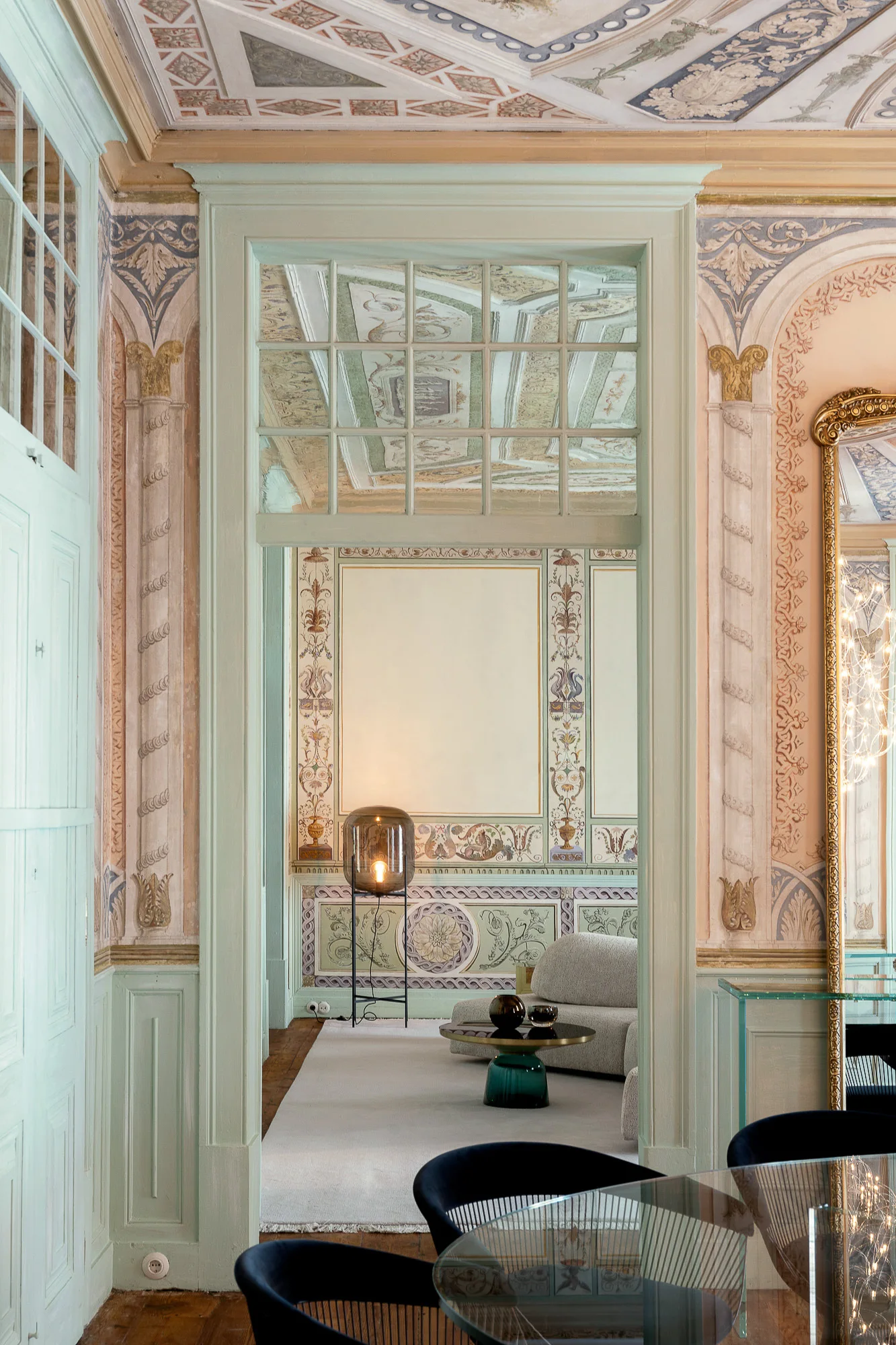 A view through a pale green painted doorway into a living room featuring ornate ceiling and wall murals, a curved sofa, and a glass-topped dining table.