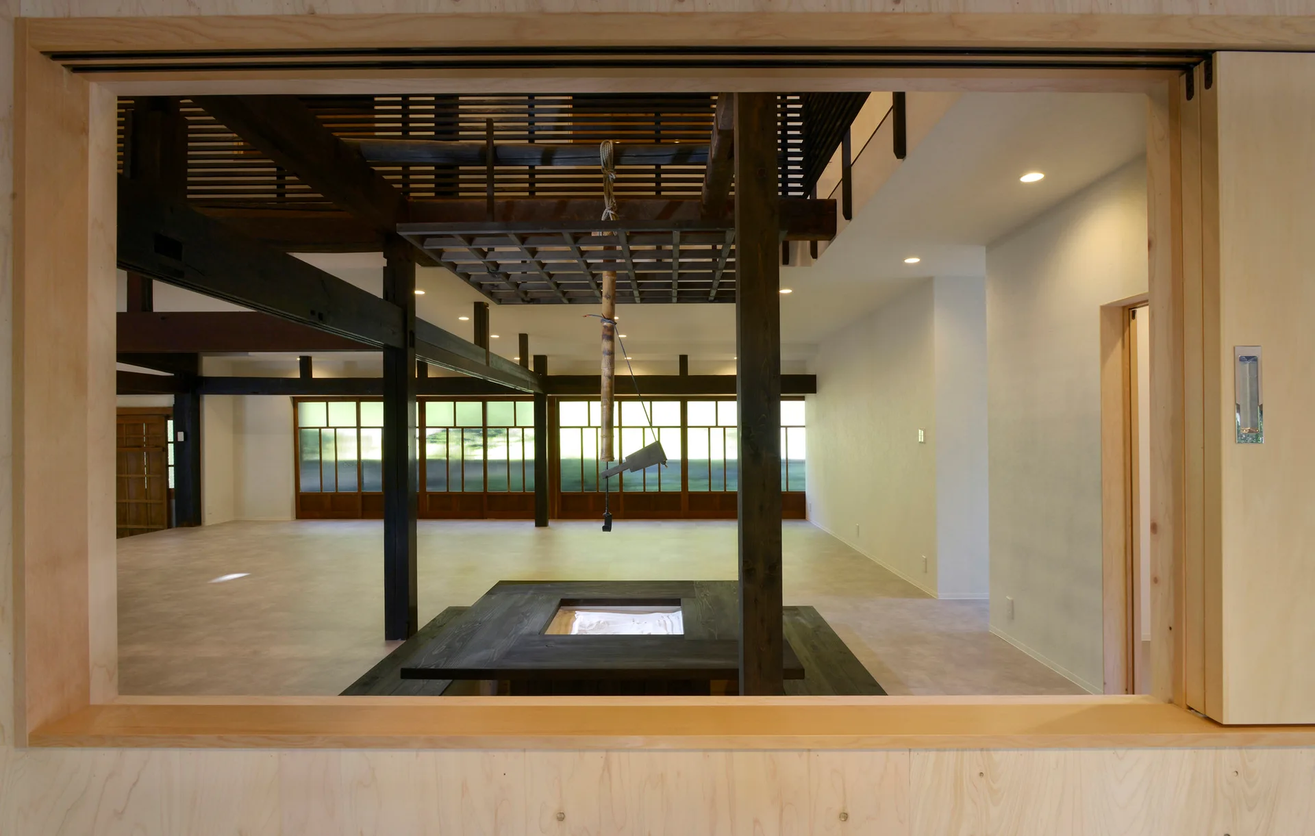 A minimalist open-plan room features a central dark wood irori hearth table, exposed dark timber beams, and grid-patterned sliding glass windows.