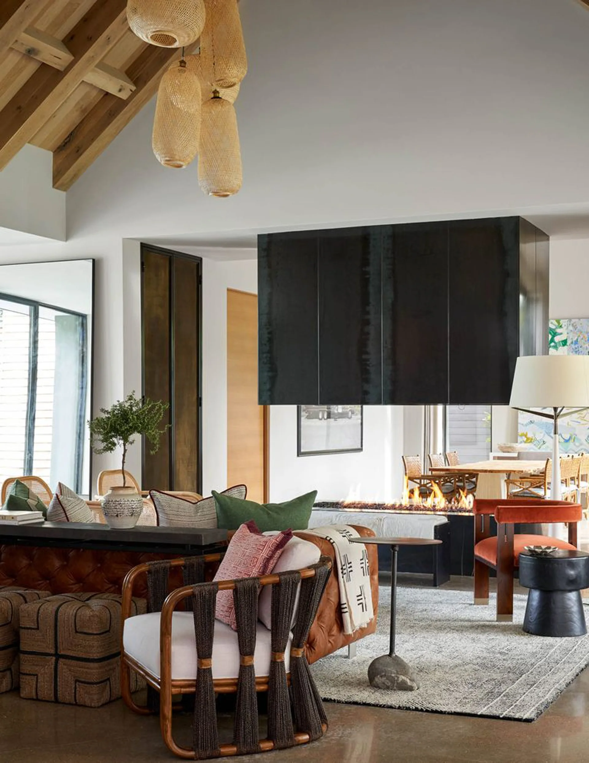 A living room features a suspended black fireplace, a tufted leather sofa, a rattan chair, and woven pendant lights under a vaulted wood ceiling.