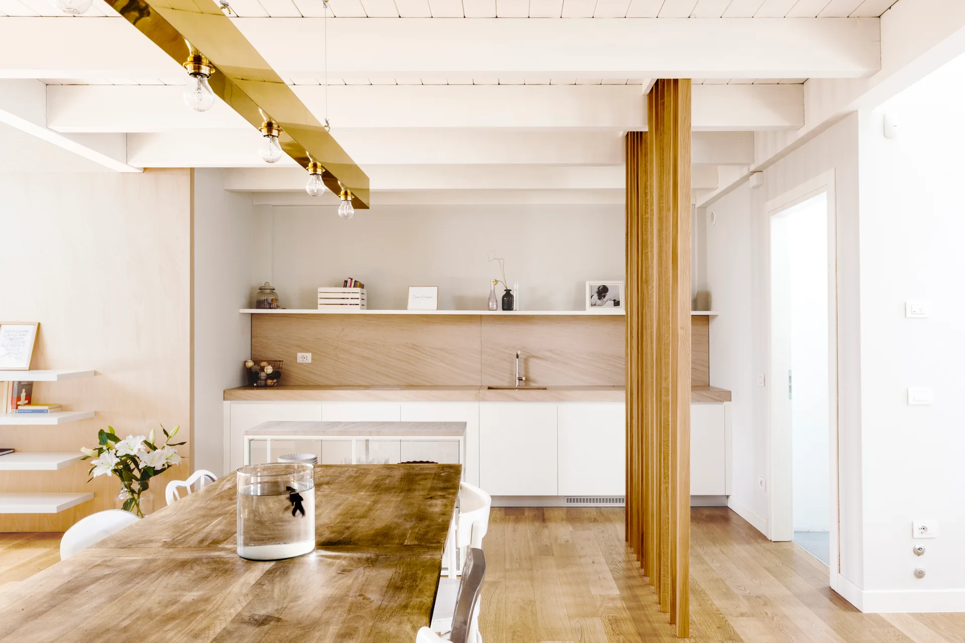 A dining area features a wooden table under a brass pendant light, adjacent to a white kitchen with a vertical wood slat partition.