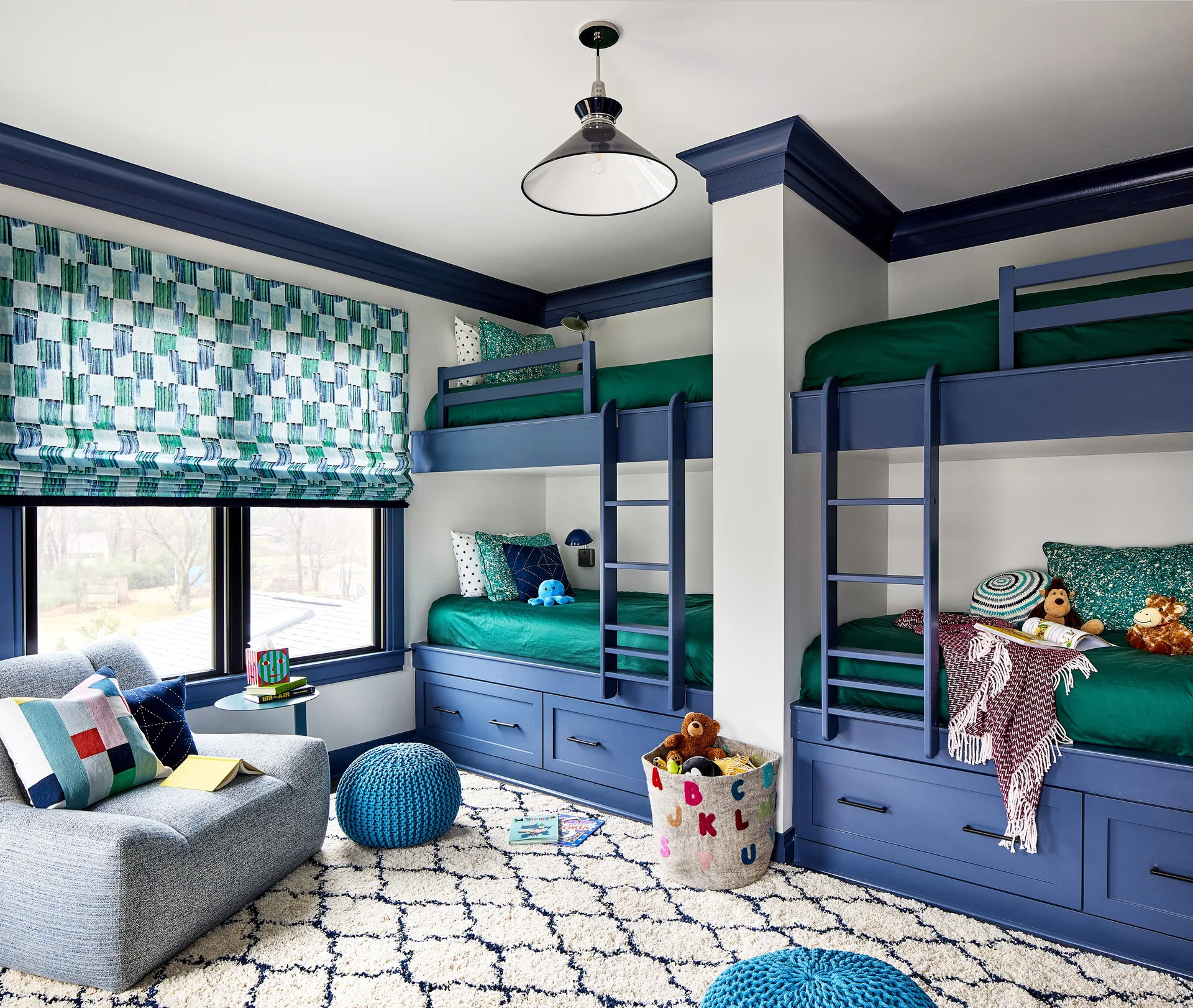 A shared children's bedroom features two sets of built-in navy blue bunk beds, emerald green bedding, a patterned window shade, and a trellis rug.