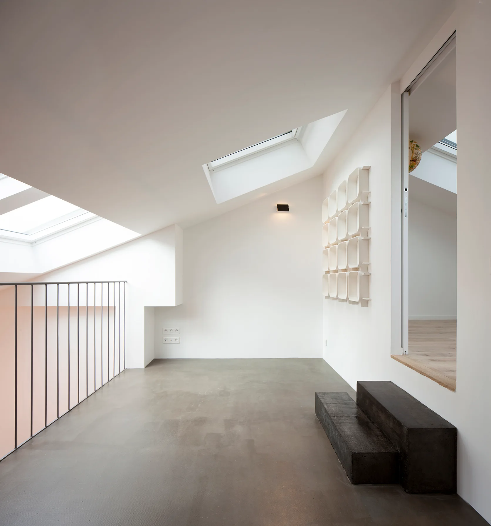A minimalist landing features slanted white ceilings with skylights, a thin metal railing, a smooth grey concrete floor, and two dark block steps.