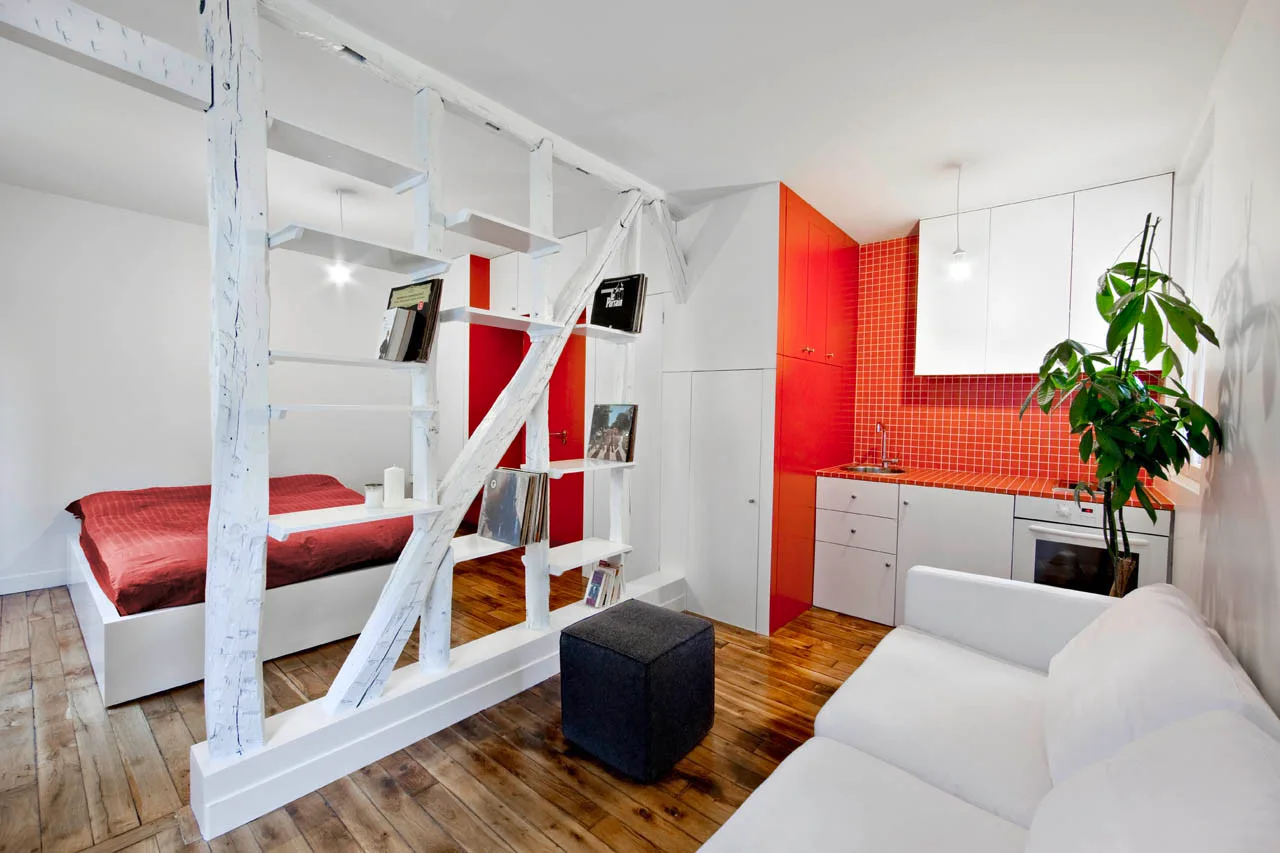 A studio apartment showing a white timber partition separating a bed from a living space and a kitchen with a vibrant red tiled backsplash.
