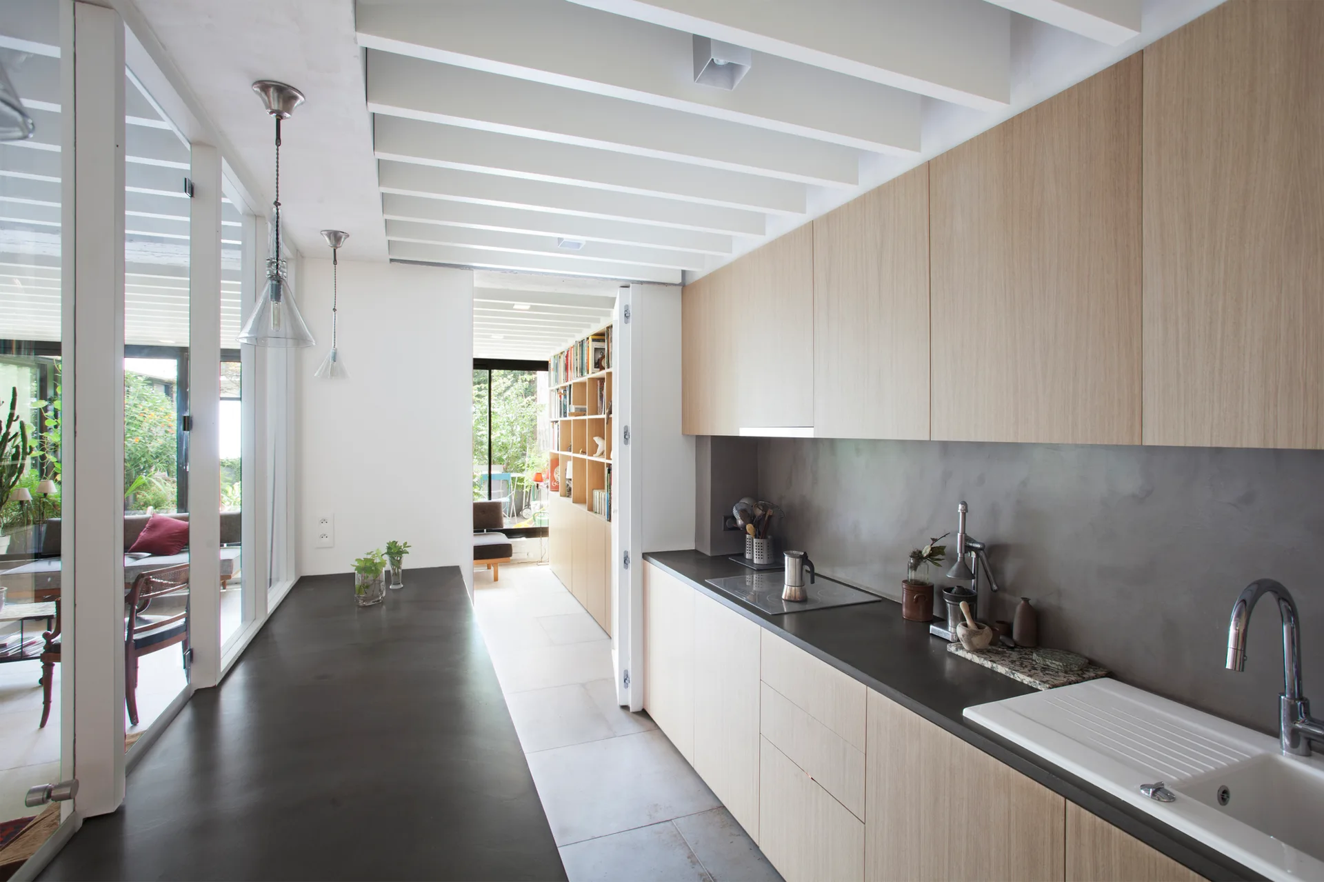 A modern galley kitchen features minimalist light wood cabinets, dark matte countertops, a grey backsplash, and an exposed white beam ceiling.