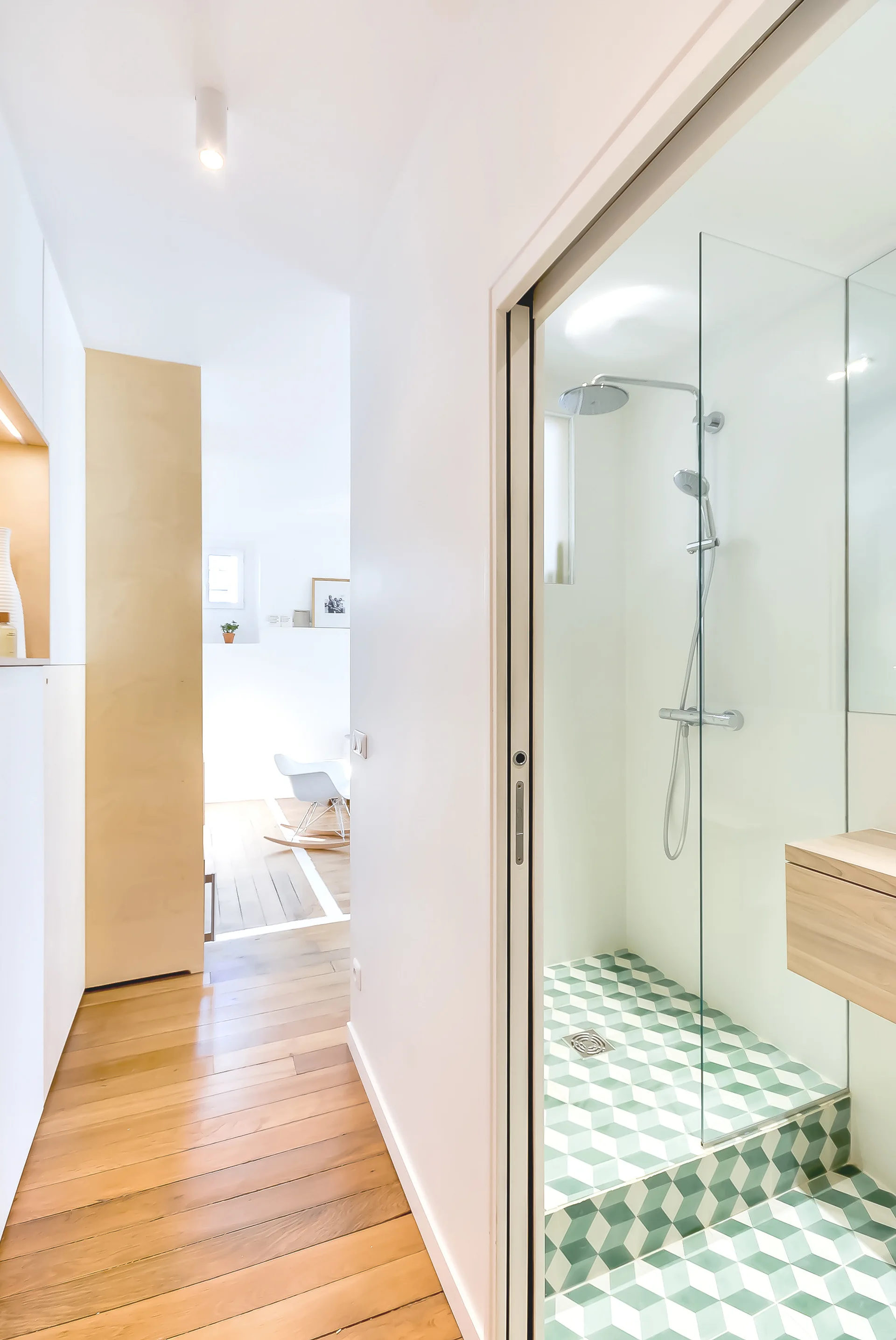 A hallway view of a bathroom featuring a glass-enclosed shower with green and white geometric floor tiles next to a floating wooden vanity.