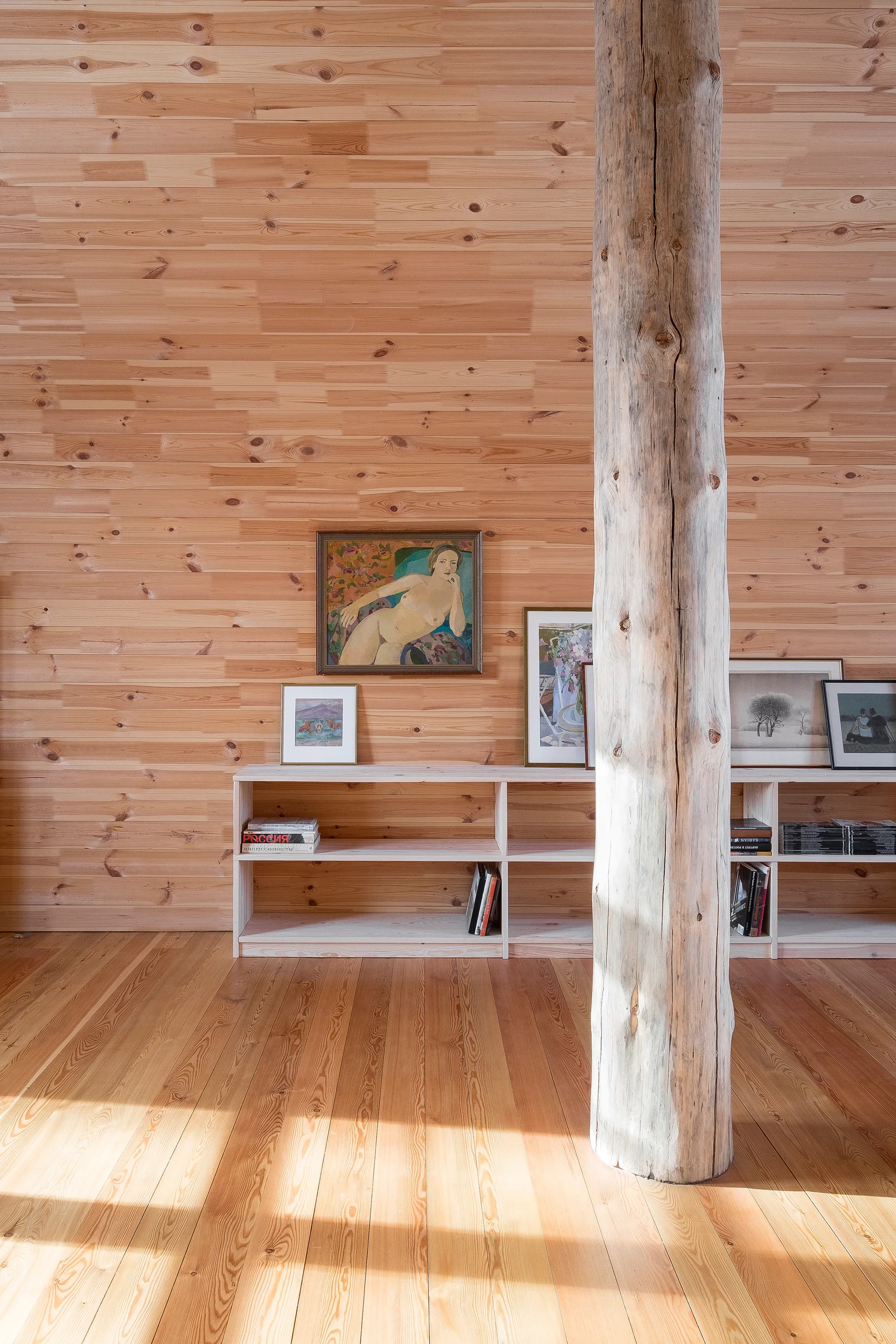 A rustic interior with horizontal knotty pine wood paneling on the walls, a raw timber pillar, and low wooden bookshelves holding framed art.