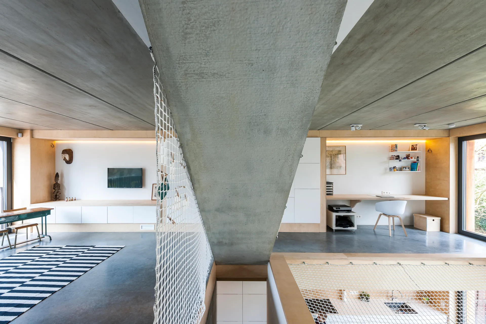 A modern loft interior featuring exposed concrete ceilings, a built-in plywood desk, white cabinetry, and a white safety net partition.