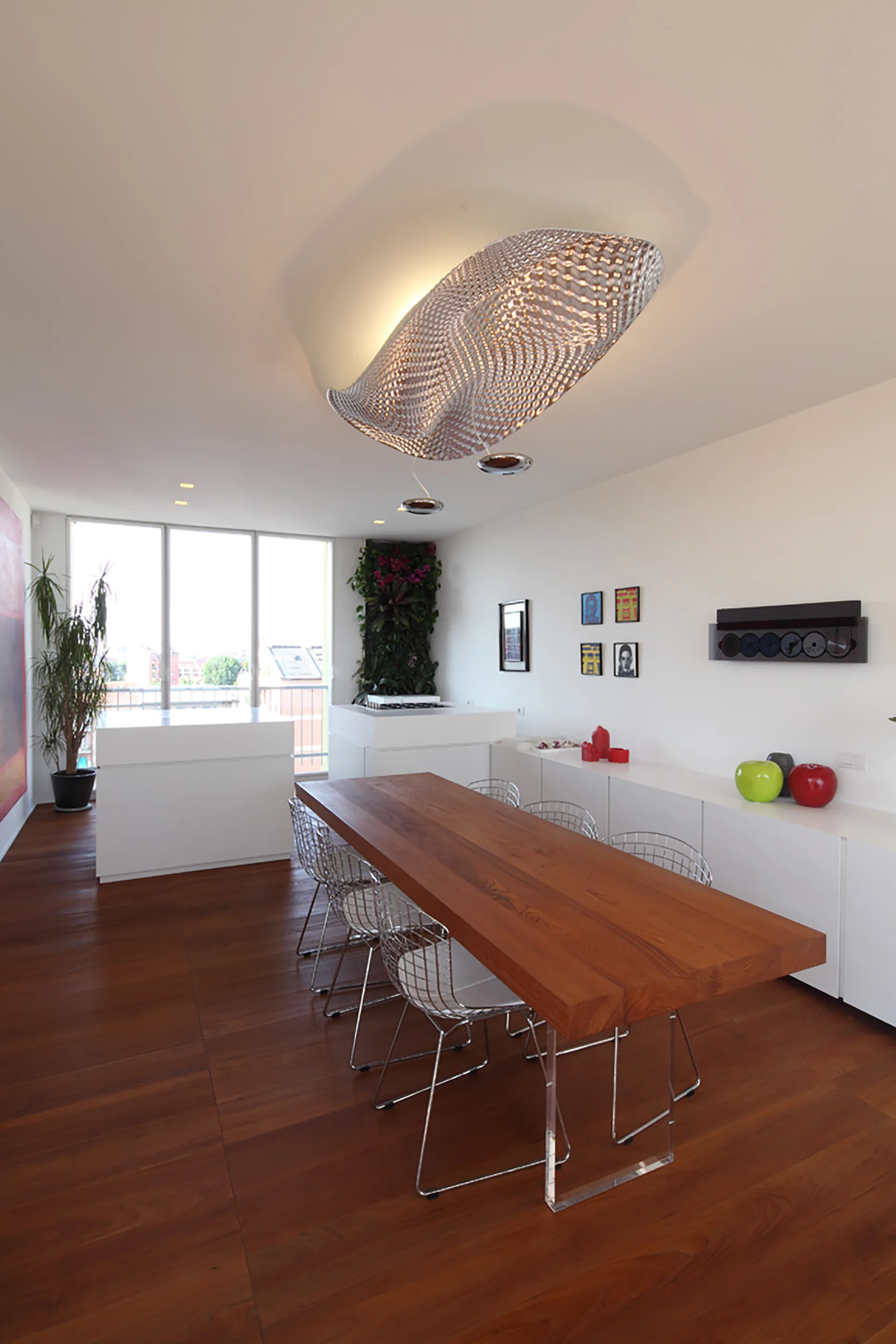 A modern dining room features a long wooden dining table, wire-mesh chairs, a sculptural ceiling light fixture, and rich wood plank flooring.