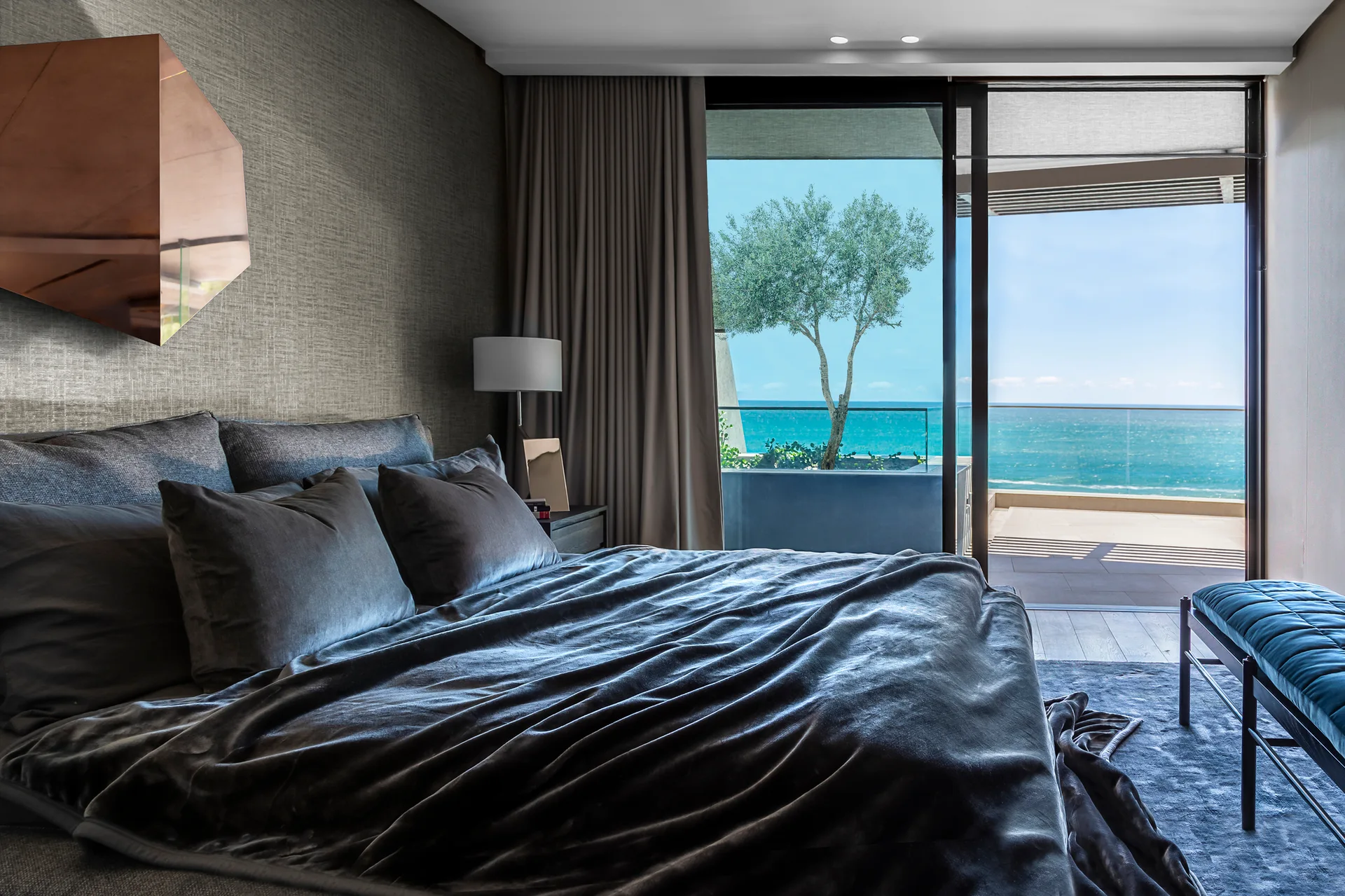 A modern charcoal gray bedroom features plush bedding, textured wallpaper with a copper mirror, and a sliding glass door opening to an ocean-facing balcony.