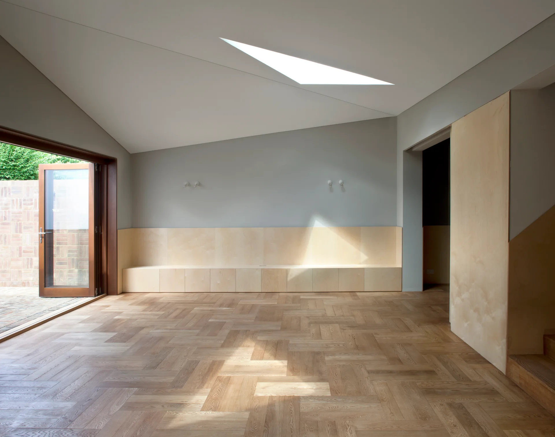 A minimalist room features light wood herringbone flooring, a low built-in storage bench against a grey wall, and an angular skylight.