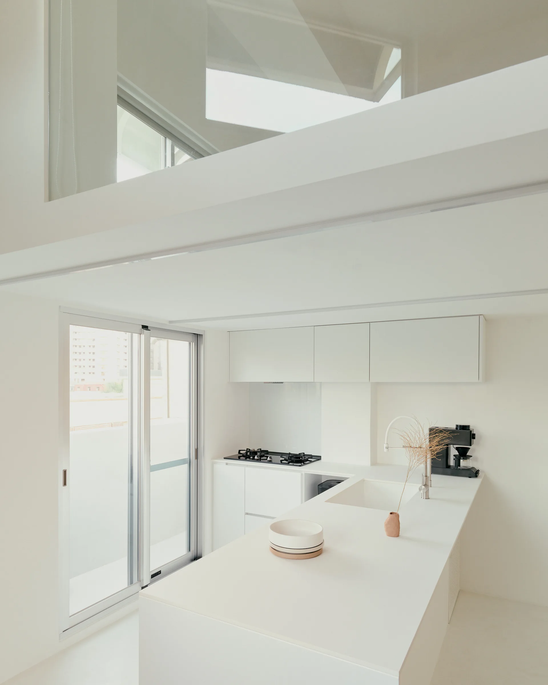 A minimalist all-white kitchen featuring a clean central island, matching cabinetry, a sliding glass door, and a mezzanine level above.