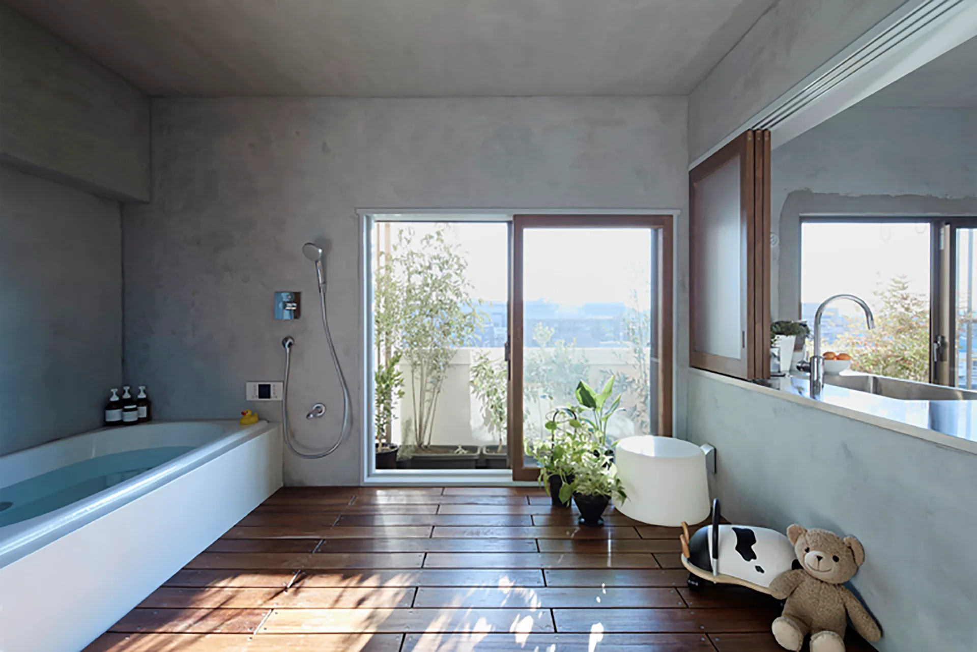 A minimalist bathroom features smooth concrete walls, a white rectangular bathtub, and wood plank flooring next to a large sliding glass door.