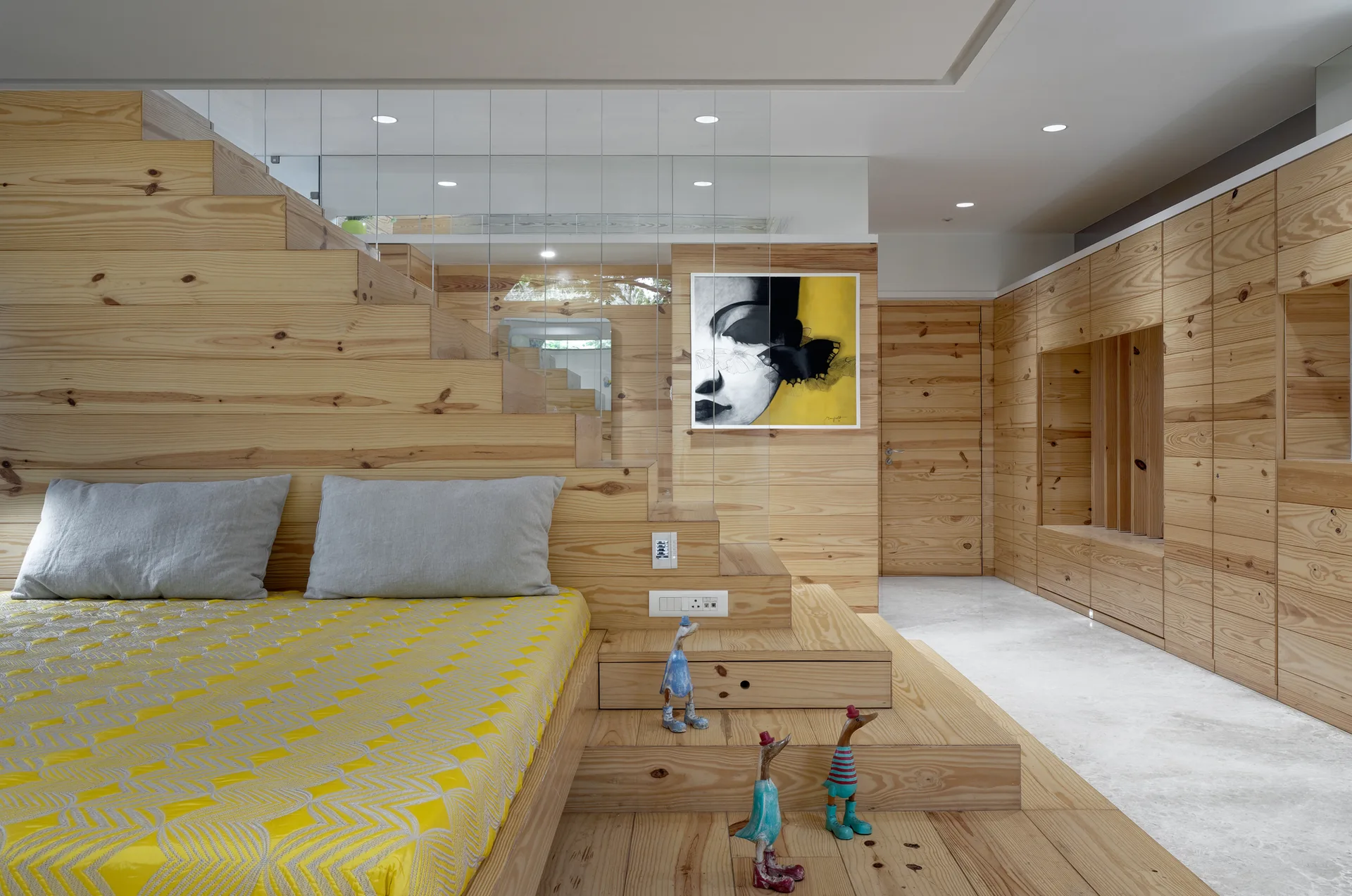 A light pine wood platform bed with yellow bedding sits next to integrated stairs, with matching wood-paneled walls and a built-in wardrobe.