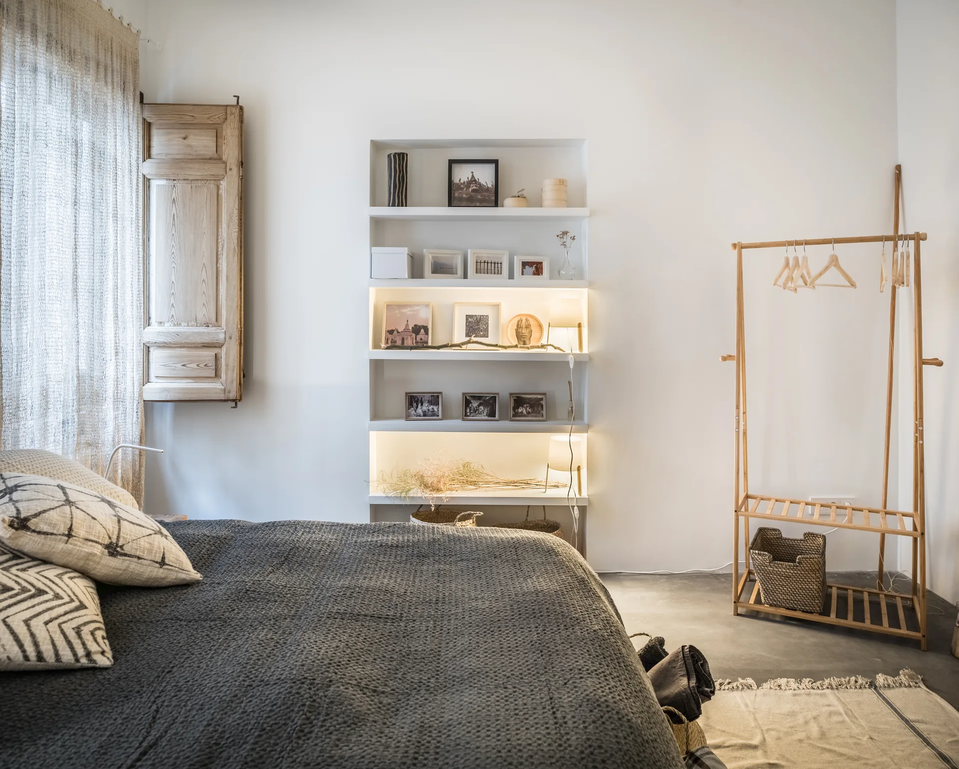 A minimalist bedroom with a gray bed, a built-in shelving unit with integrated lights, a wood window shutter, and a wooden clothing rack.