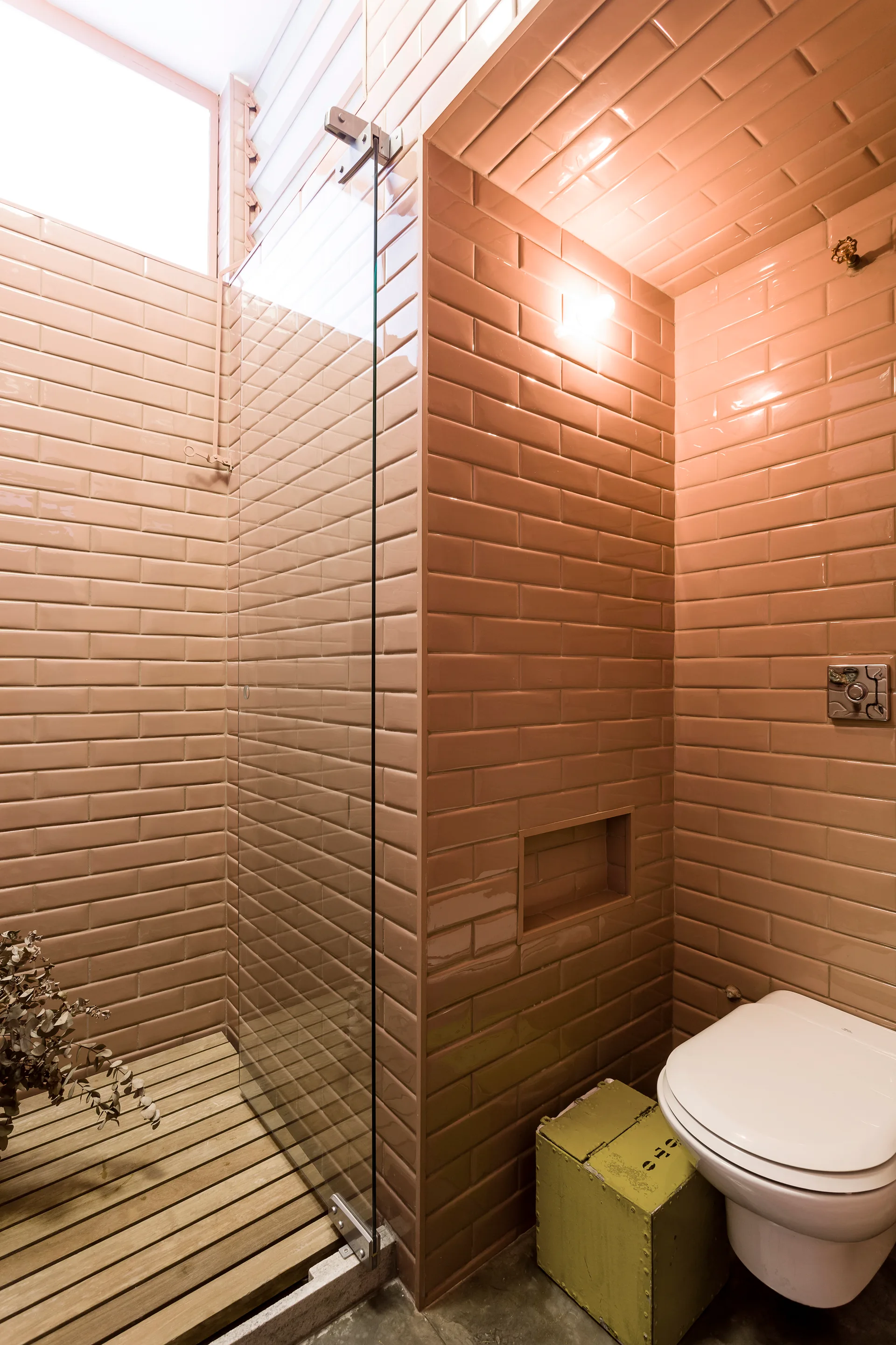 A modern bathroom featuring walls wrapped in glossy terracotta subway tiles, a glass shower partition, a slatted wood floor, and a white toilet.
