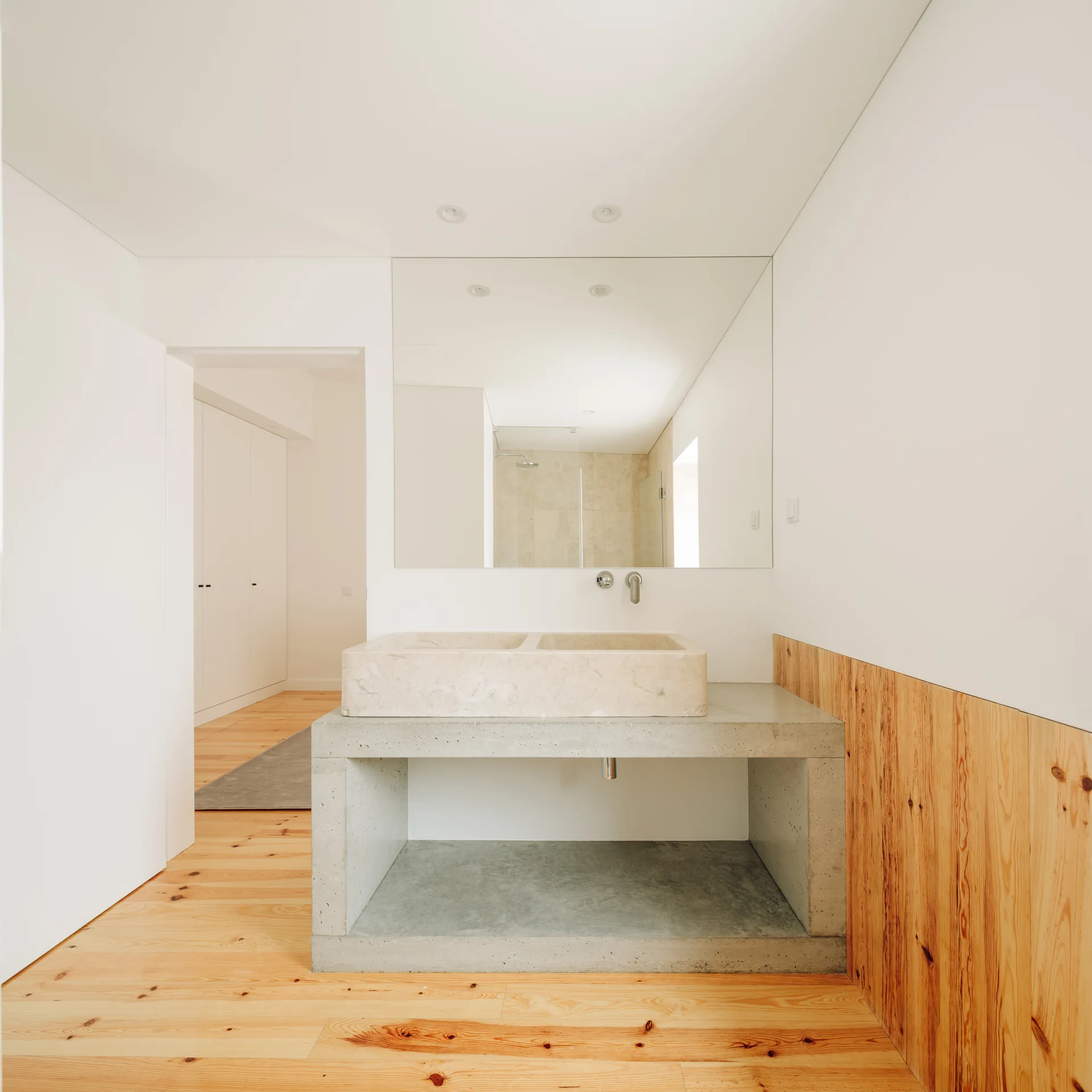 A minimalist bathroom featuring a concrete vanity block with a light stone basin sink, natural wood floors, and a large unframed wall mirror.