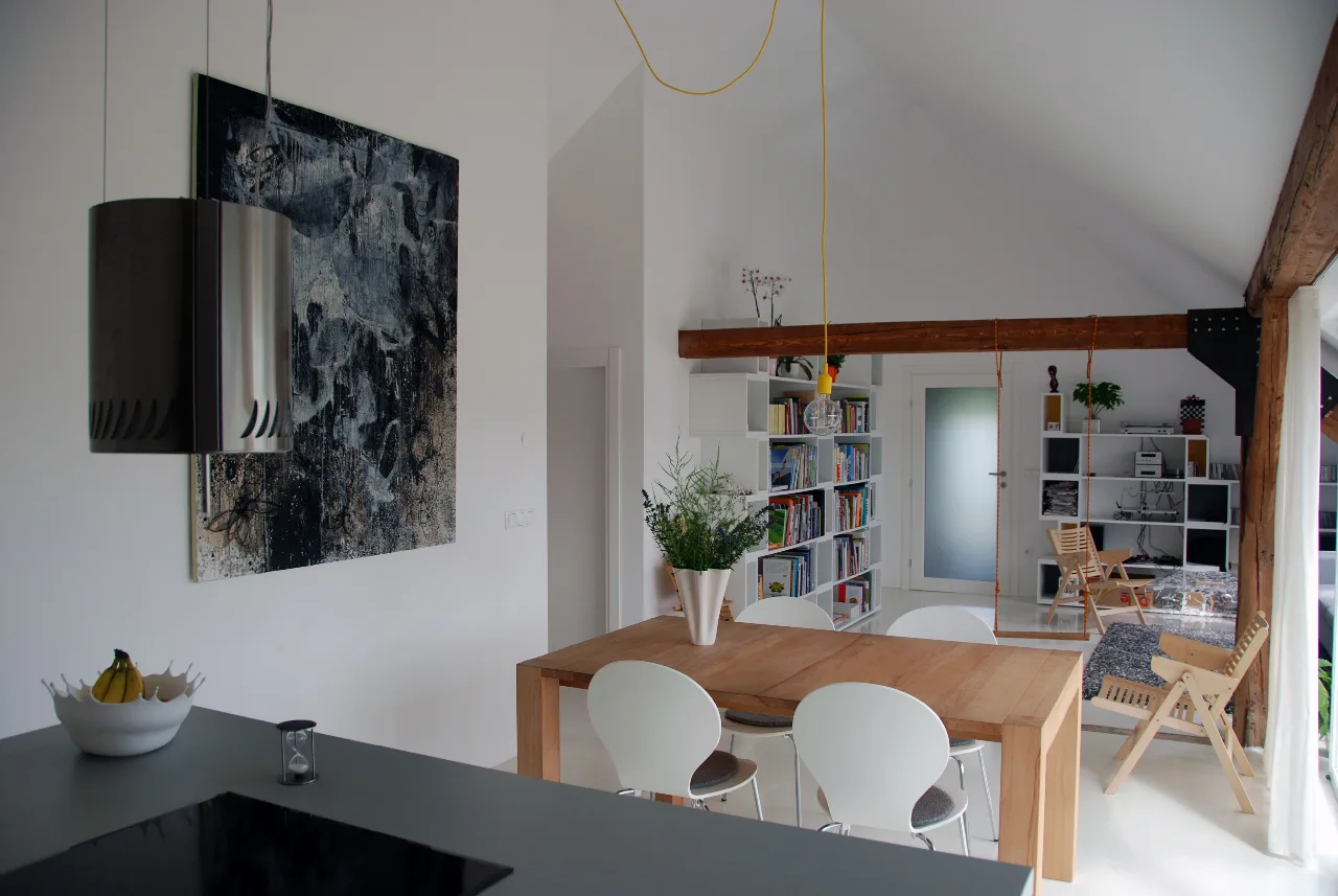 A dining area with a wooden table, white chairs, and a large abstract painting under a vaulted white ceiling with an exposed beam.