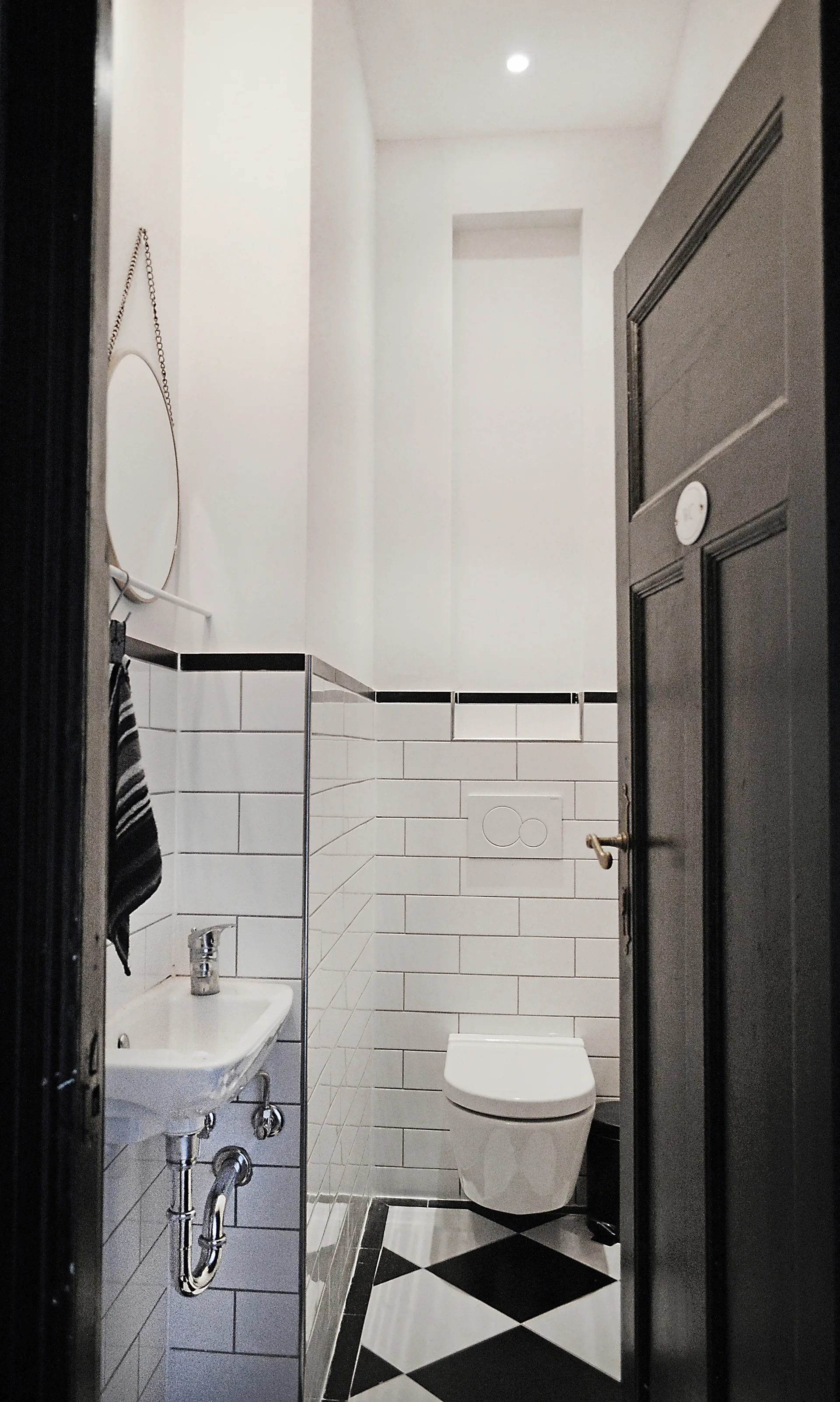 A narrow powder room features white subway tile walls, a wall-mounted toilet, and a black and white checkerboard tile floor viewed through a dark door.