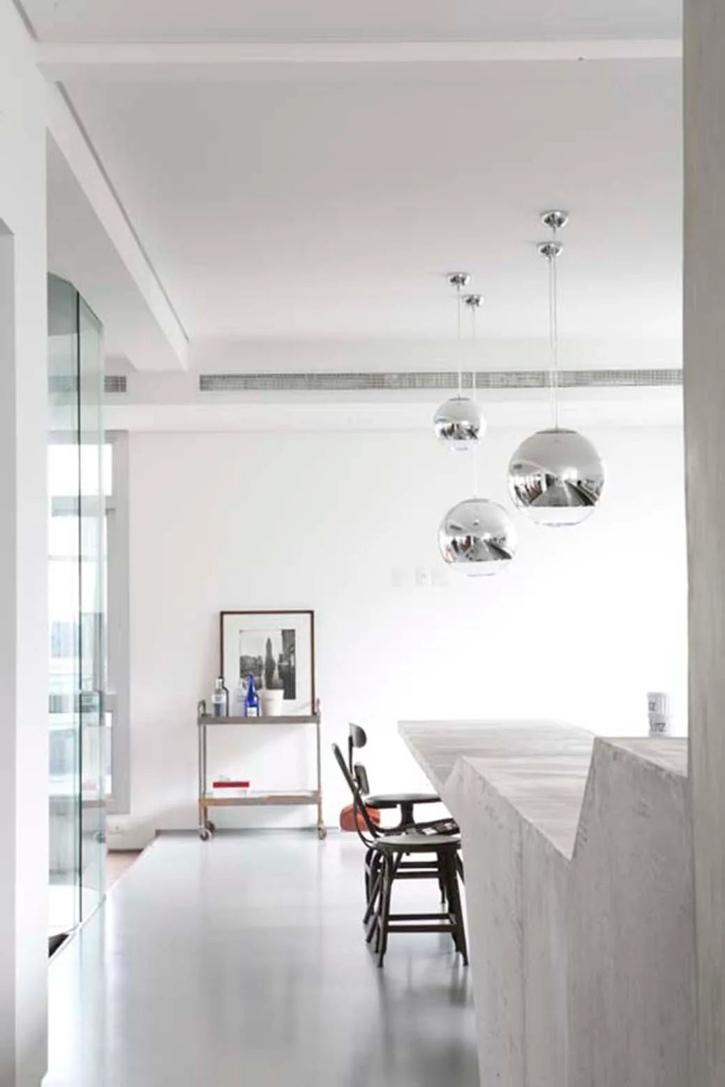 A minimalist kitchen area featuring a textured concrete island, dark bar stools, and three reflective spherical pendant lights.