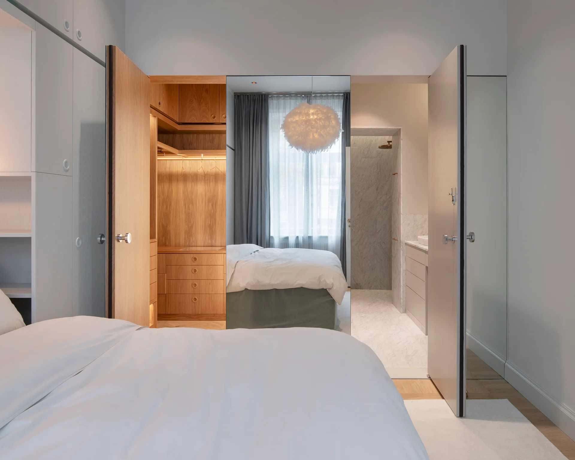 An open-plan bedroom suite featuring a built-in oak wardrobe with warm lighting and a doorway leading into a marble bathroom.