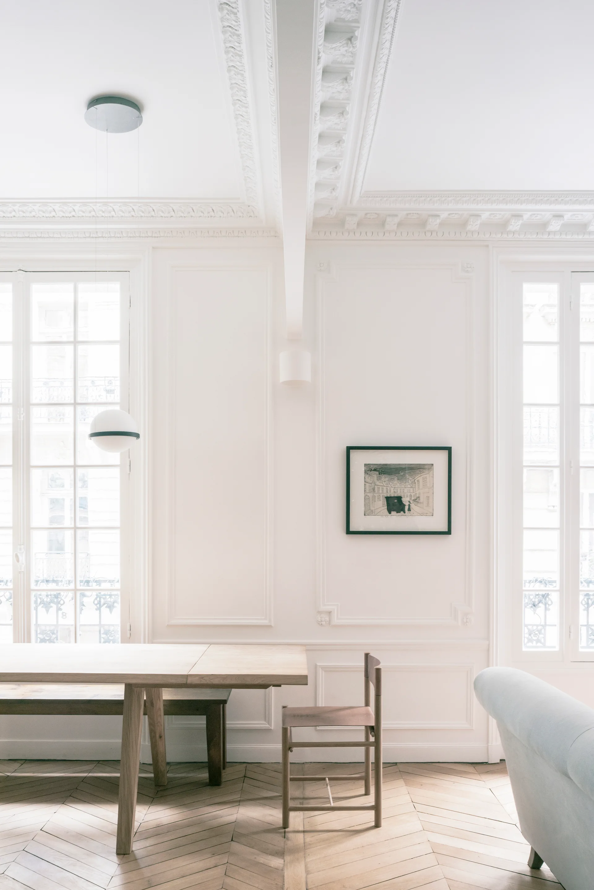 A light-filled room featuring white paneled walls with detailed moldings, chevron-patterned wood floors, a simple wooden dining table, and tall French windows.