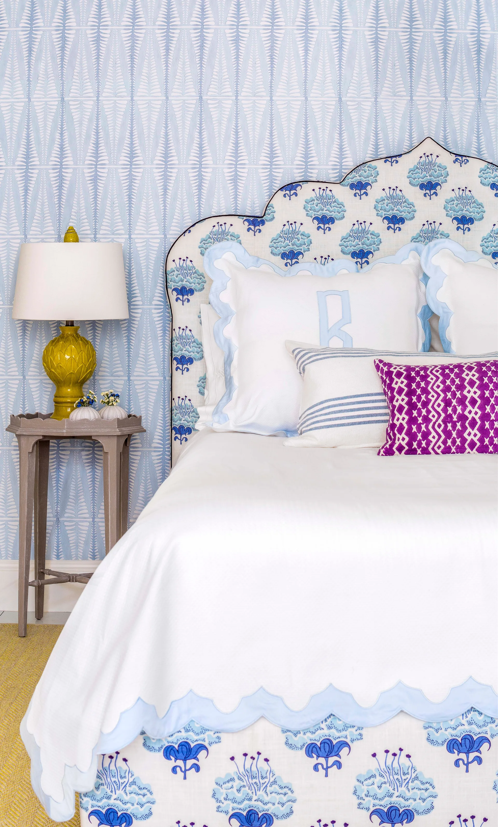 A bedroom features a bed with a blue floral upholstered headboard, white monogrammed pillows, a chartreuse table lamp, and light blue patterned wallpaper.