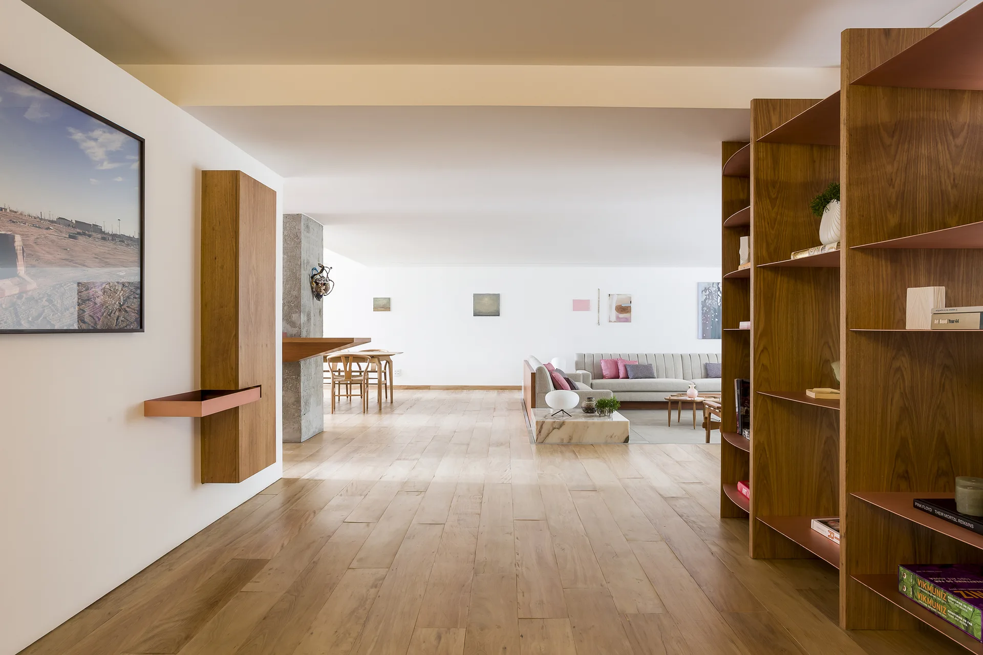 An open-plan apartment interior features hardwood flooring, tall wooden bookcases, a distant dining area, and a neutral sofa in the living space.
