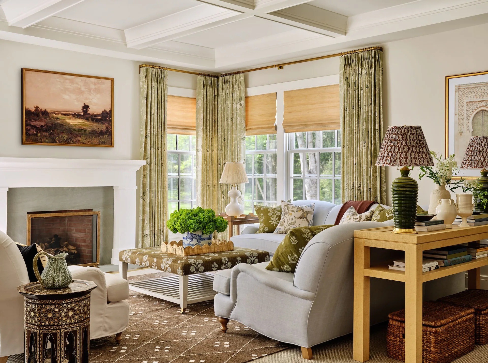 A traditional living room features a white coffered ceiling, a fireplace, light blue sofas with green floral pillows, and patterned drapery.