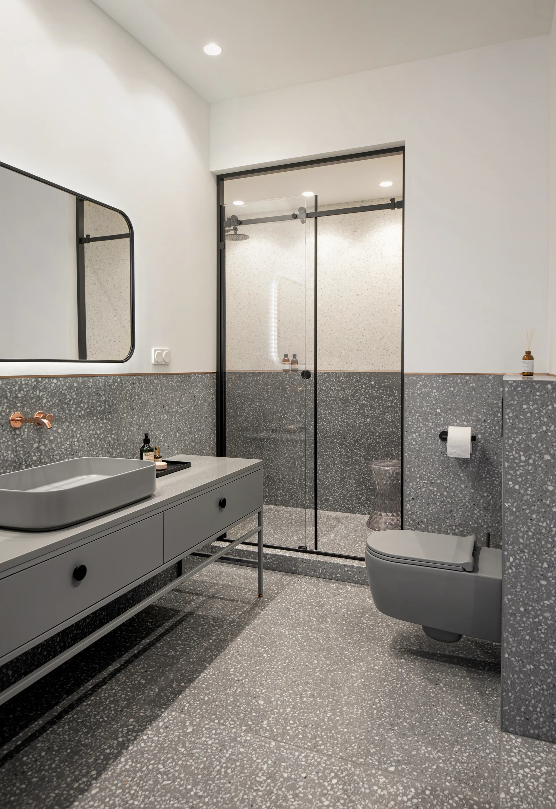 A modern grey bathroom featuring a matching terrazzo floor and half-wall, a floating grey vanity with a vessel sink, and a wall-hung toilet.