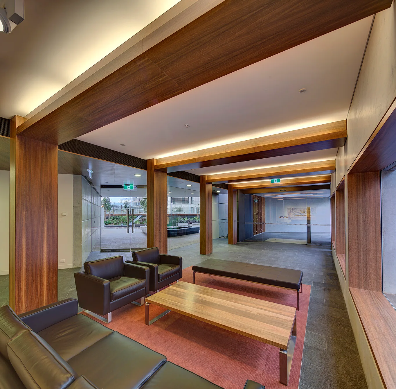 Black leather furniture sits on a red rug in a modern lobby featuring a wood-beamed ceiling, stone floor, and large wood-framed windows.