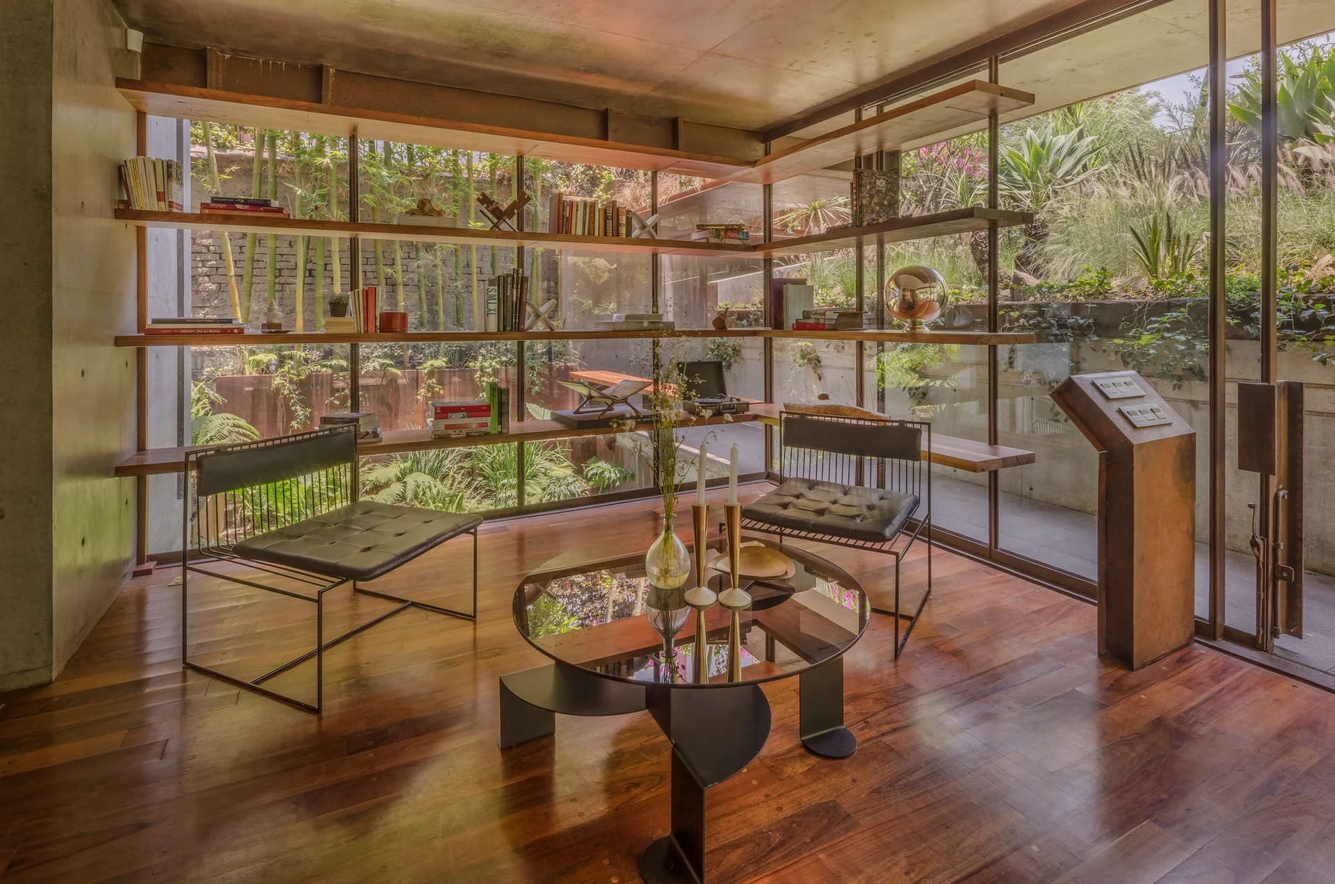 A modernist living area features floor-to-ceiling glass walls, a concrete ceiling, wrap-around wooden bookshelves, and two black leather armchairs.