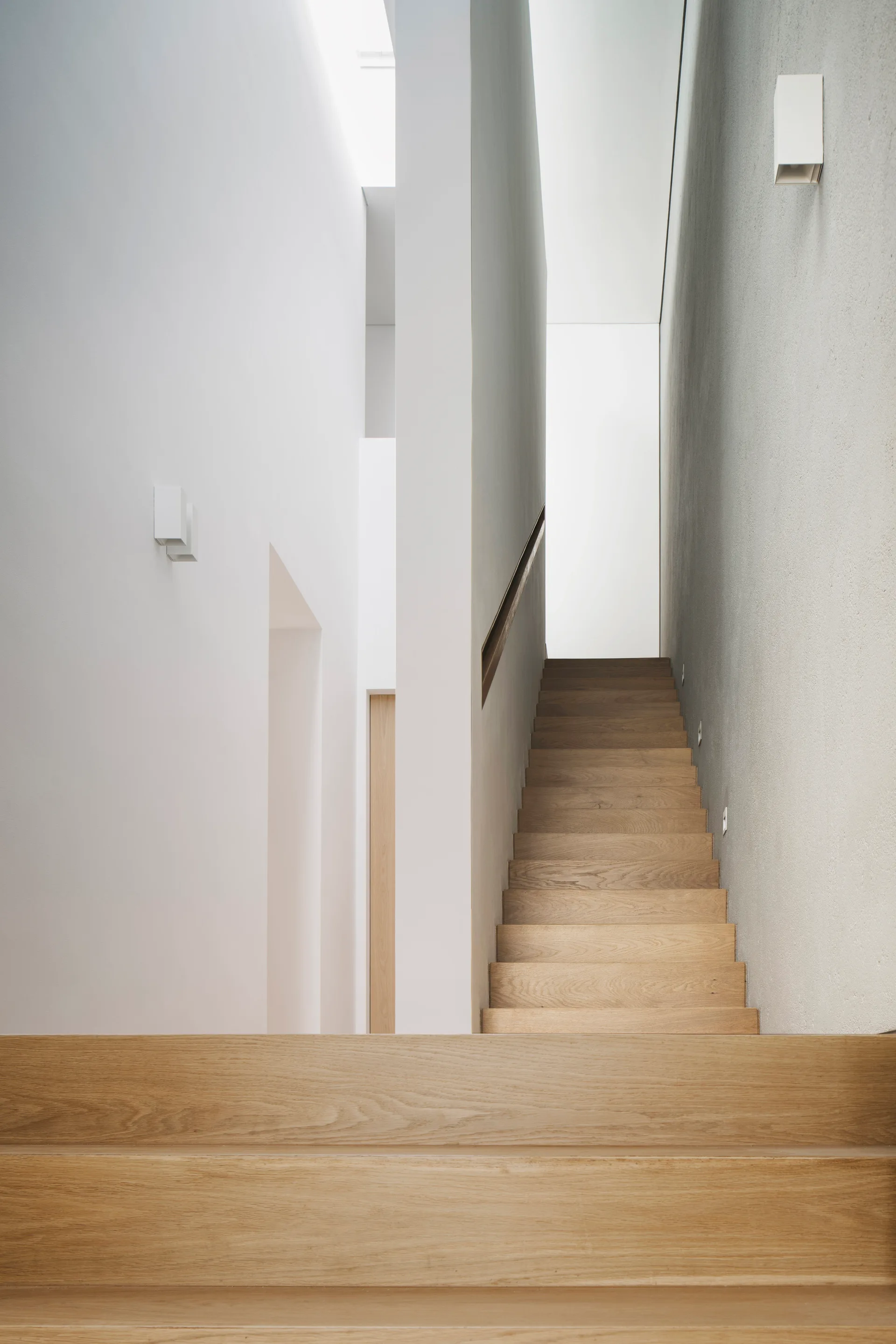 A minimalist modern staircase features light oak wood treads between smooth white walls with a recessed handrail and simple white sconces.