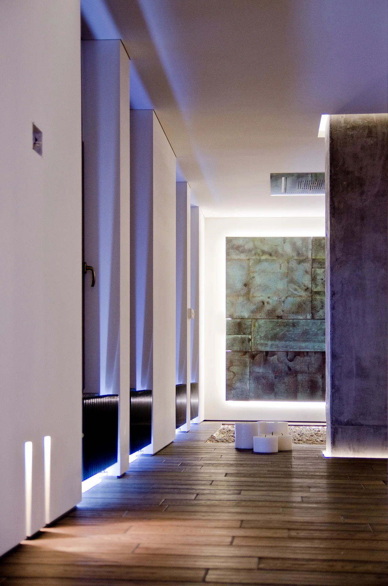 A minimalist hallway featuring dark wood plank floors, white architectural wall columns with baseboard LED lights, and a backlit patinated metal art panel.