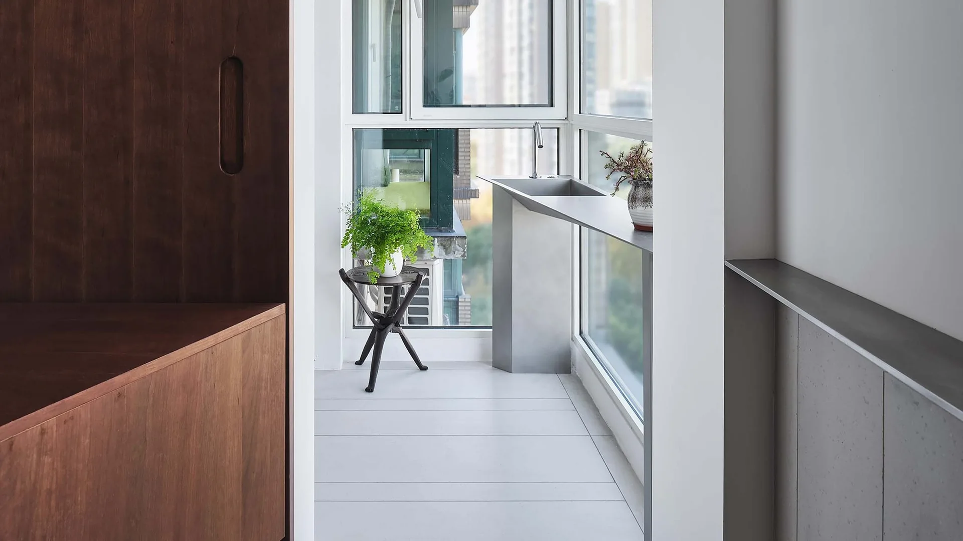 A modern minimalist balcony features a slim metal sink console against large windows, light grey tile flooring, and dark wood built-in cabinetry.