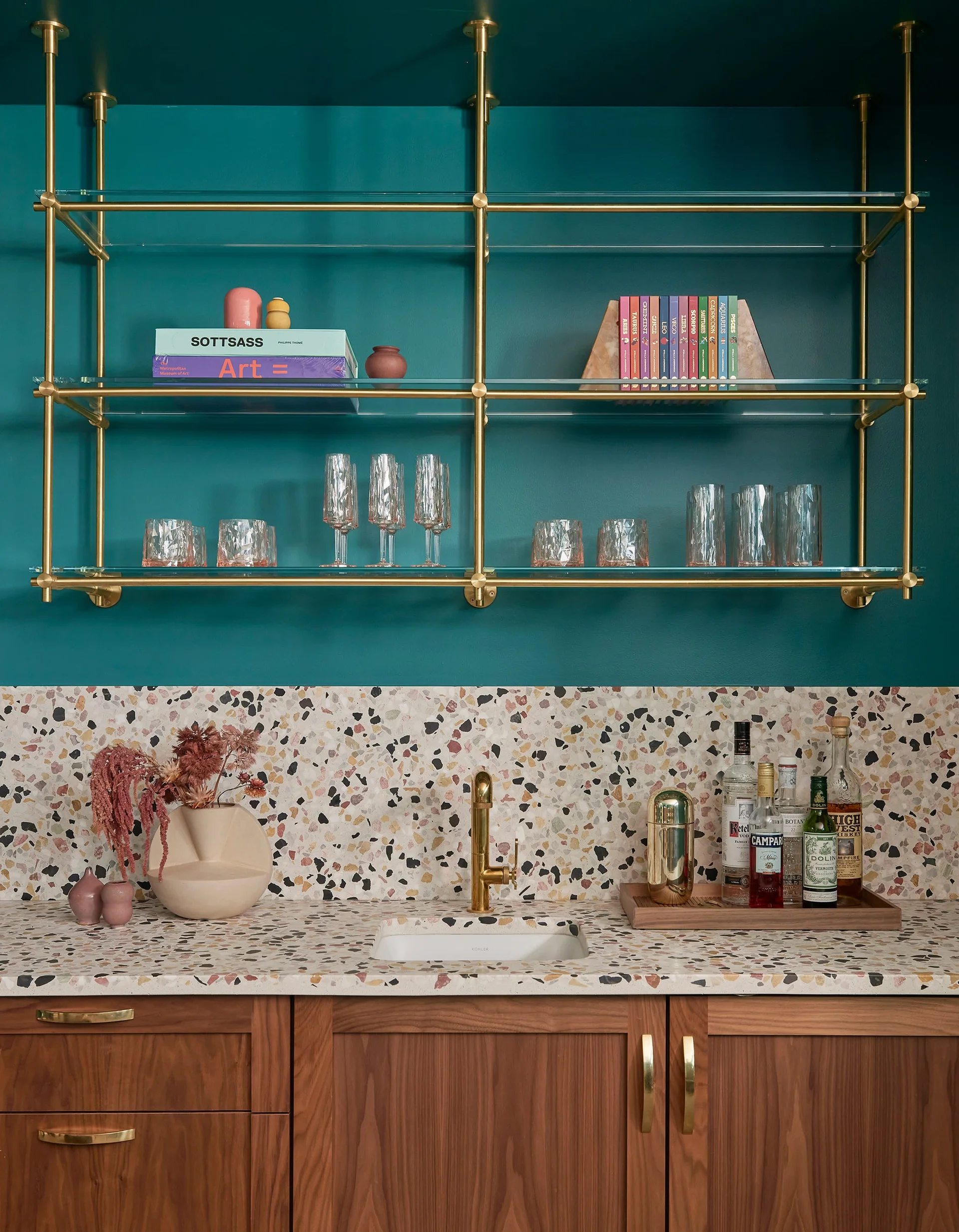 A wet bar features wood cabinets, a colorful terrazzo countertop and backsplash, brass faucet, and glass shelving against a teal wall.