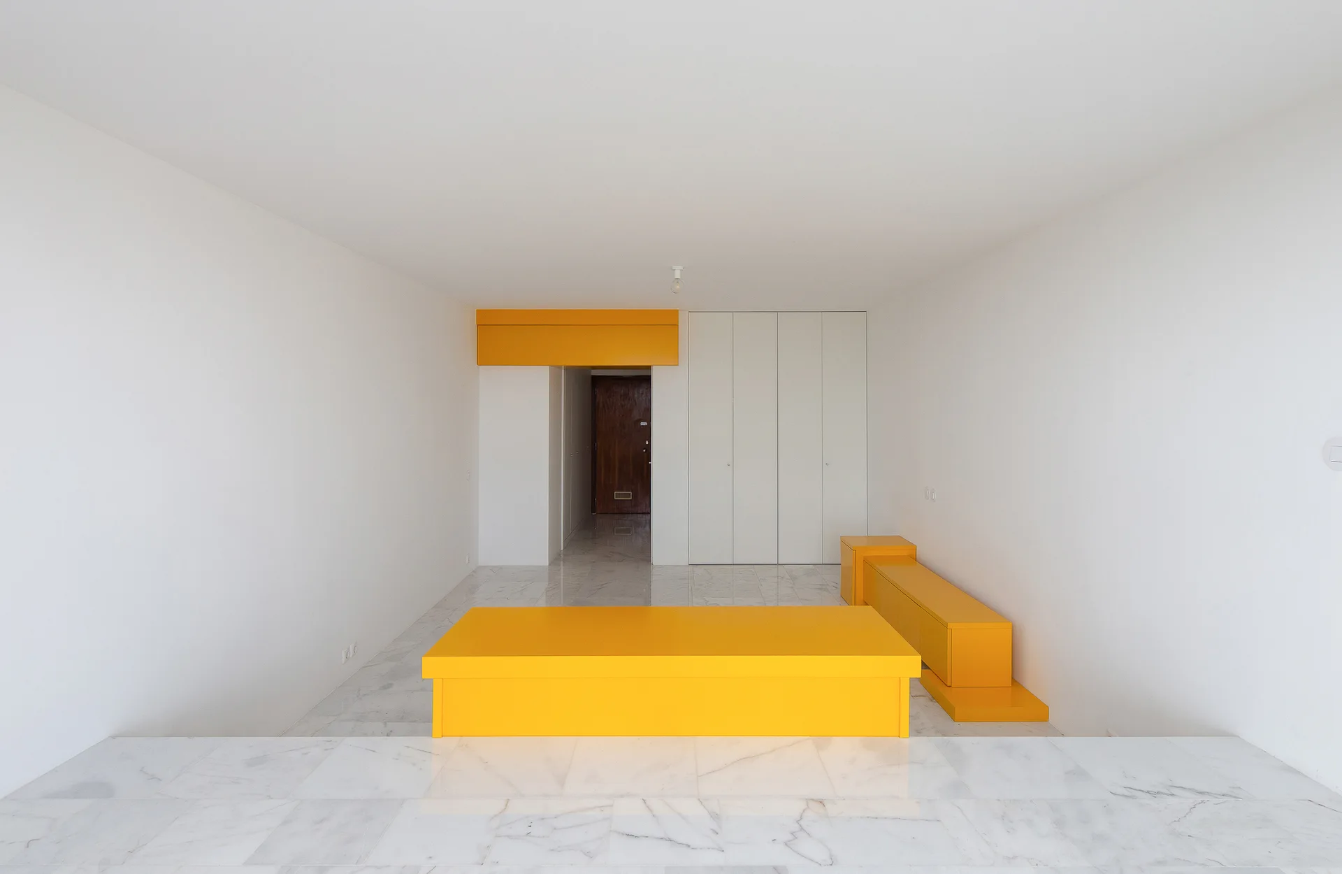 A minimalist white room with polished marble flooring features custom geometric yellow furniture including a low block bench and wall console.