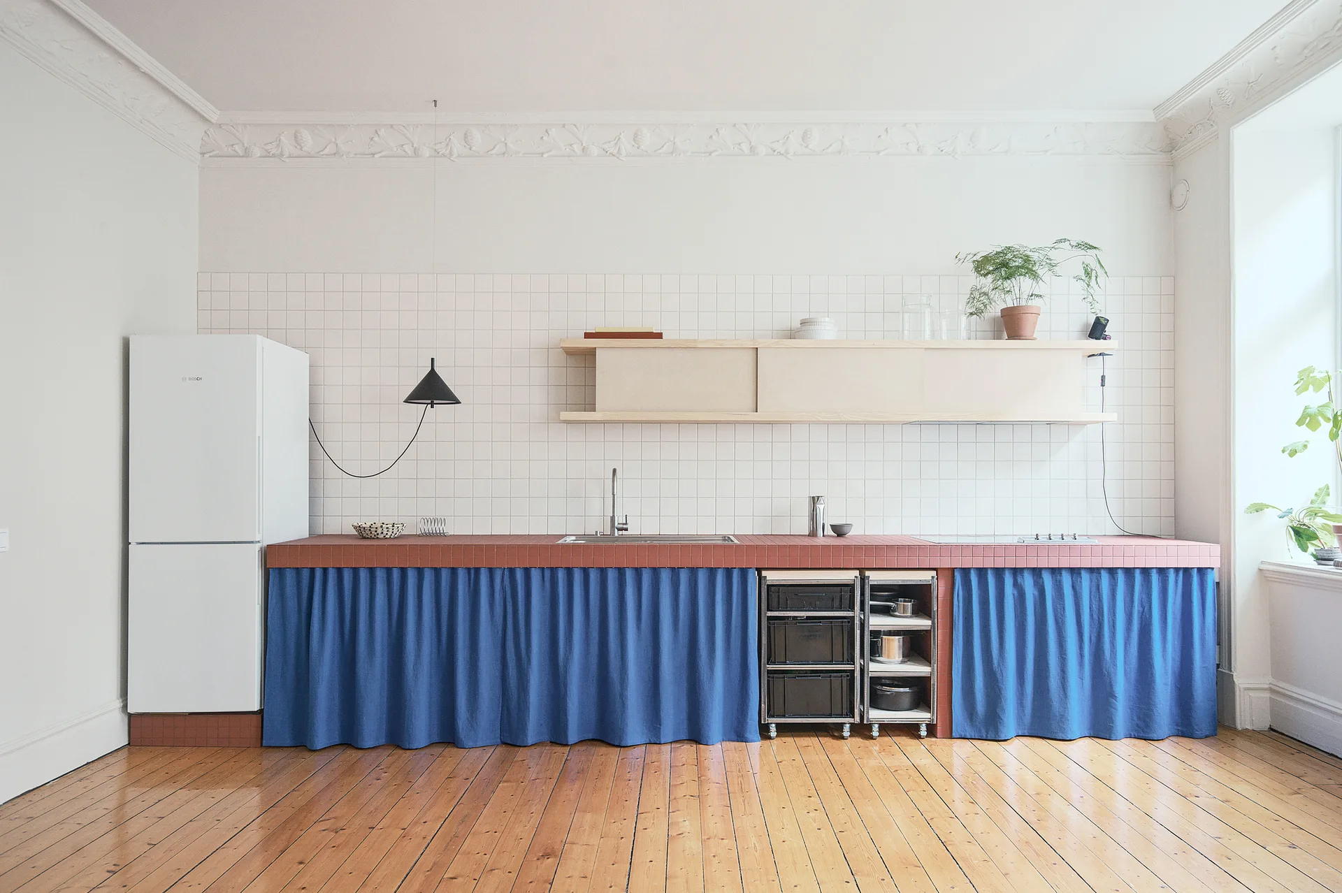 A minimalist kitchen features a terracotta tiled countertop with blue linen curtains underneath, a white tile backsplash, and light wood flooring.
