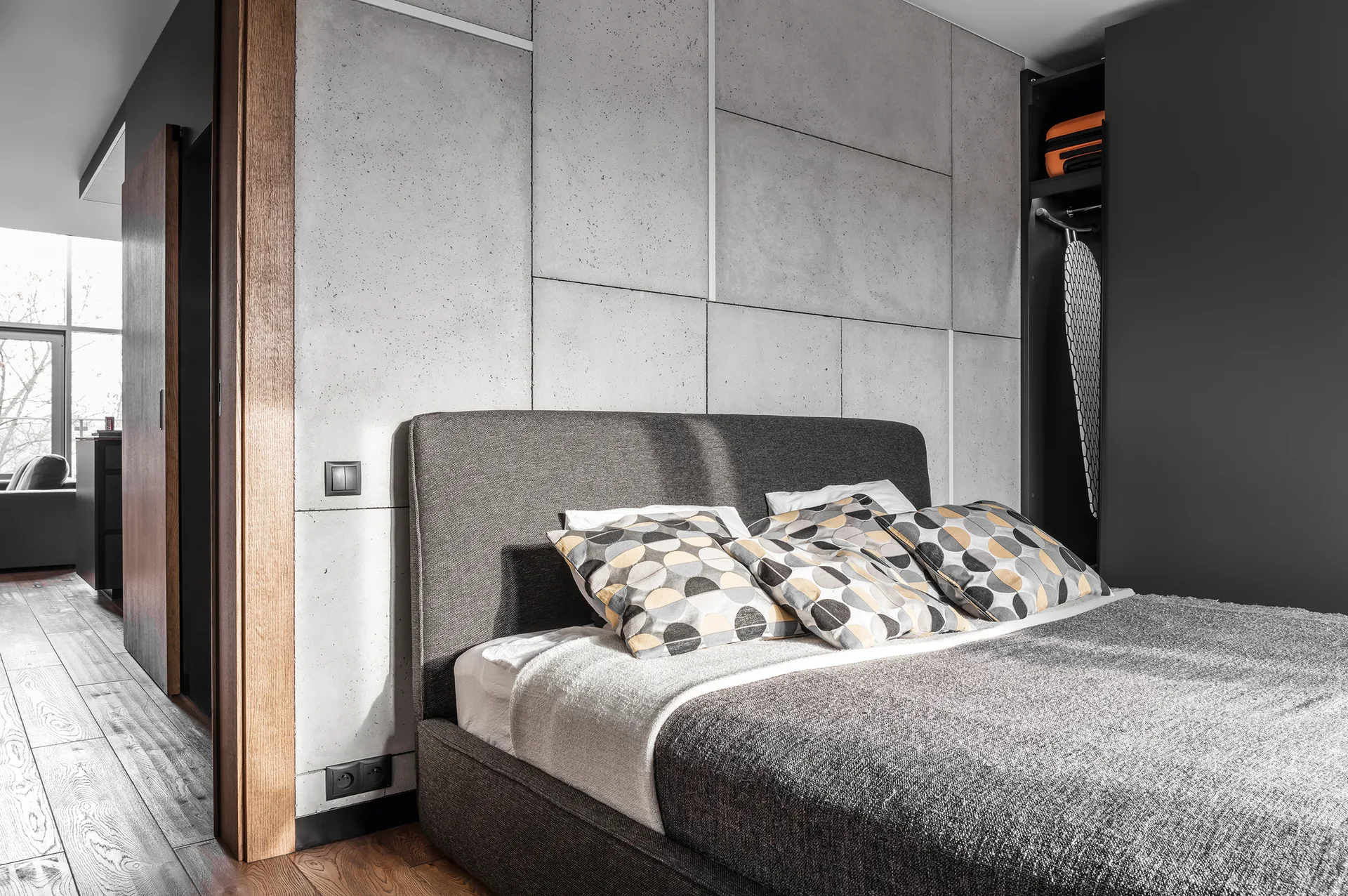 A grey upholstered bed is set against a textured concrete panel accent wall in an industrial style bedroom with wood flooring.
