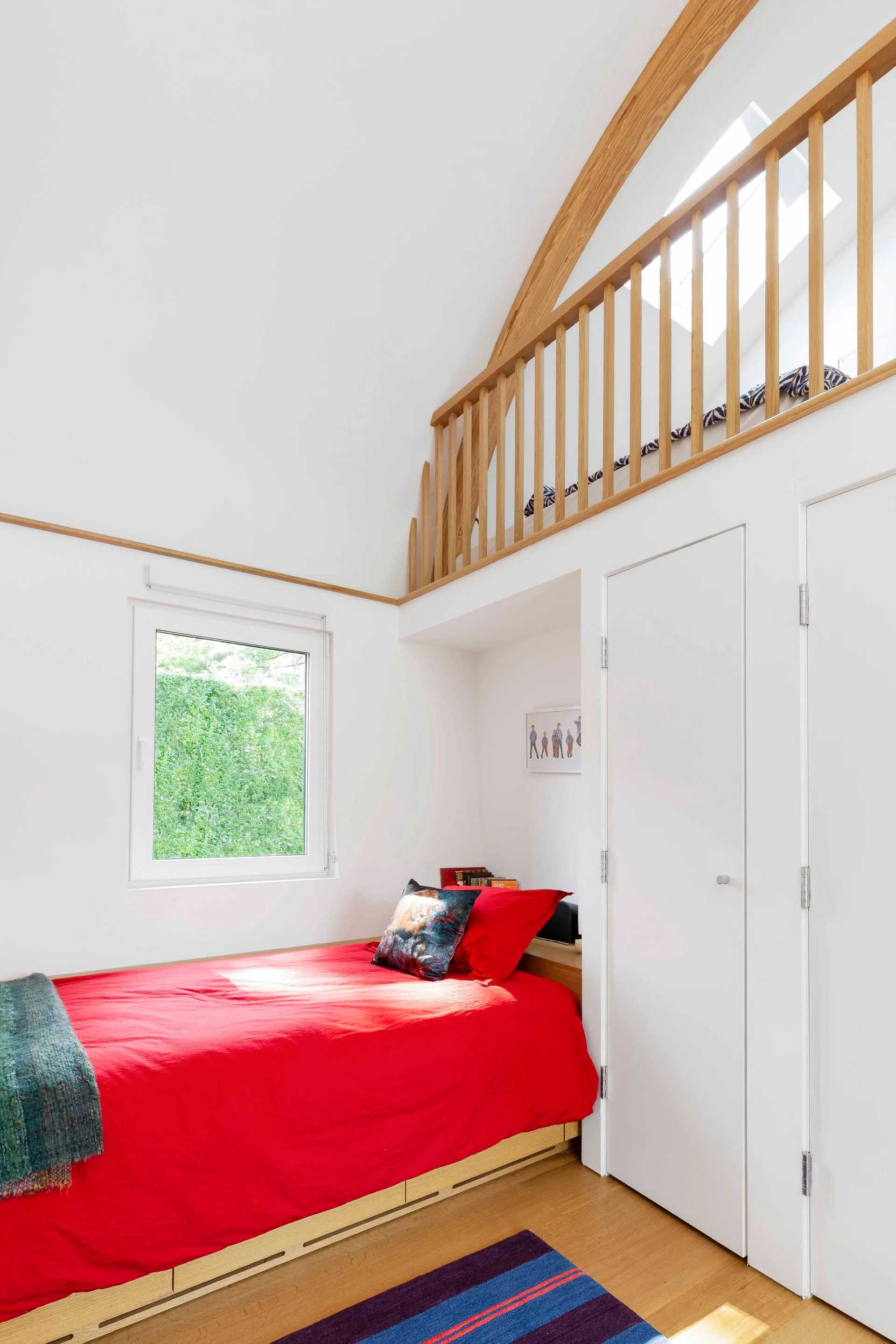 A bright white bedroom features a bed with vibrant red bedding beneath a wooden mezzanine loft with vertical wood slats.