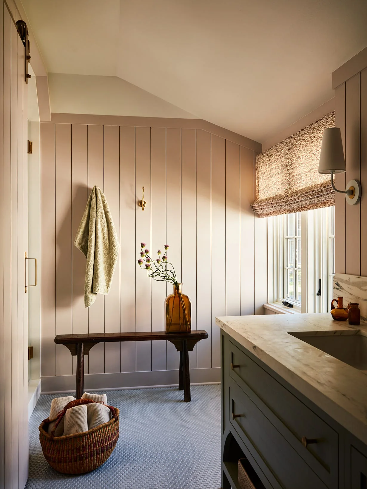 A bathroom featuring pale pink vertical wood-paneled walls, a sage green vanity with a marble countertop, a small dark wood bench, and penny floor tiles.
