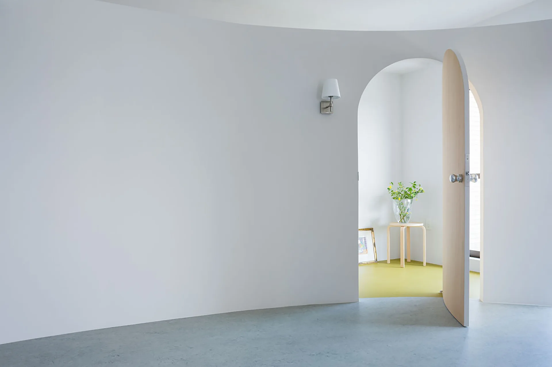 A minimalist hallway features a white curved wall and an open arched wood door revealing a room with yellow flooring and a wooden stool.