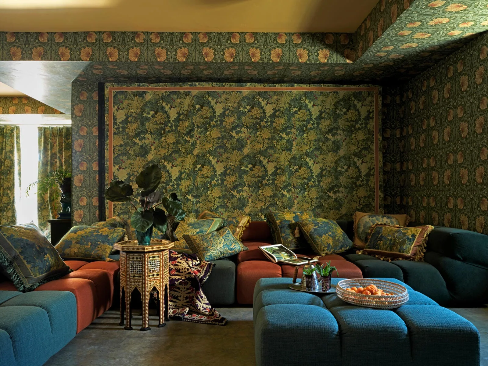 A low-profile modular sofa in teal and rust sits in a room with matching floral wallpaper on the walls and ceiling, accompanied by a blue ottoman.