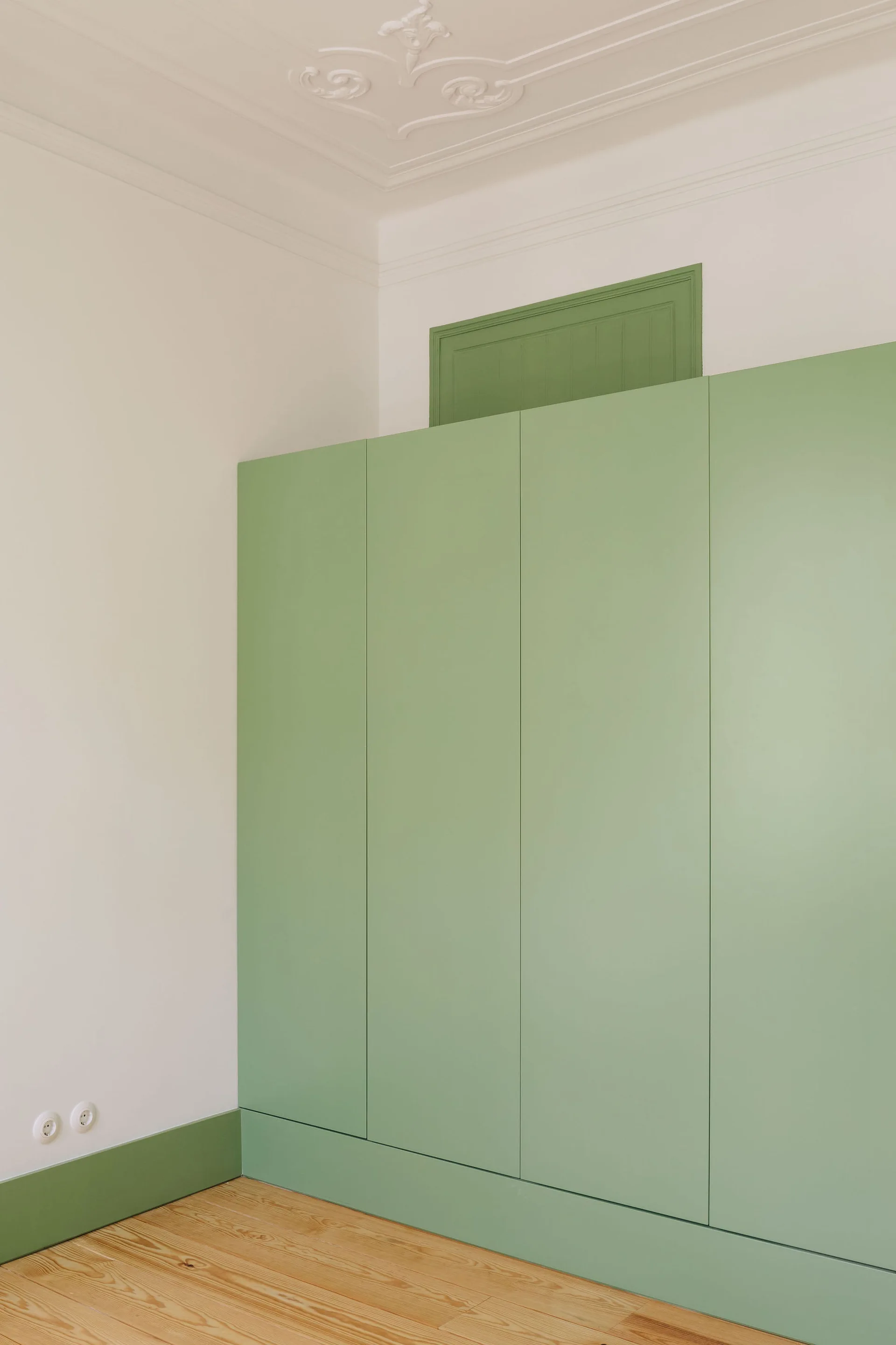A minimalist sage green built-in wardrobe stands against a white wall below an ornate plaster ceiling medallion in a room with wood floors.