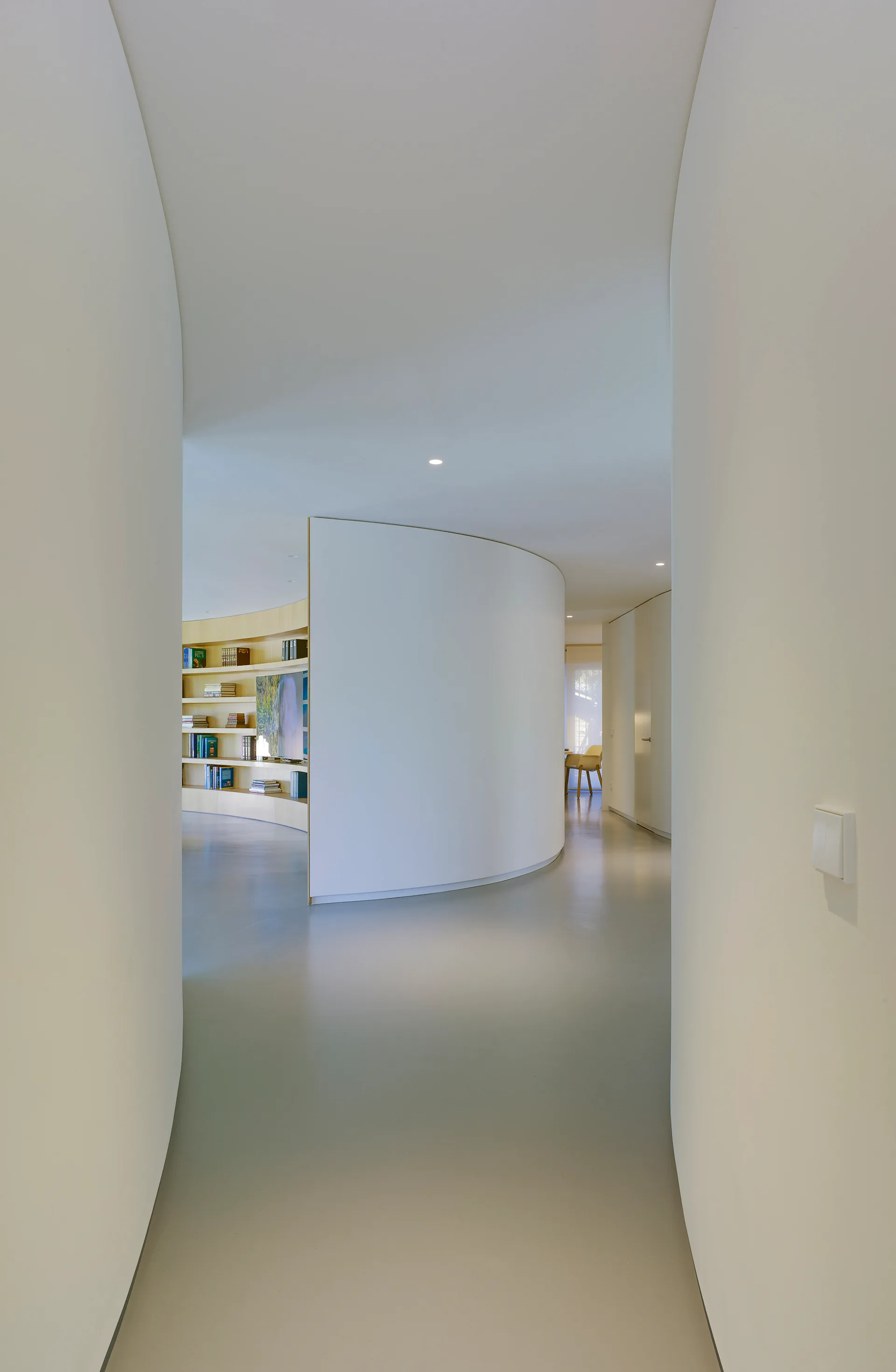 A smooth minimalist hallway with curved white walls leading past a central partition to a light wood built-in bookcase on a gray floor.