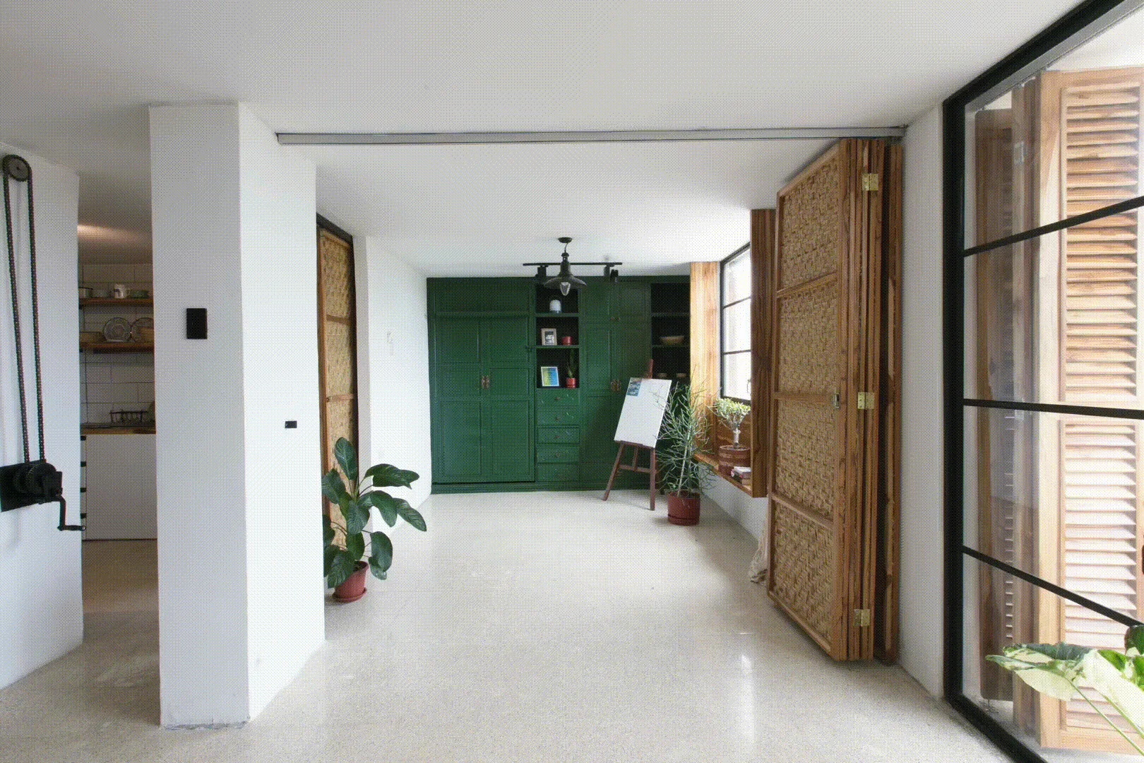 An open living space features white walls, terrazzo flooring, a large green storage cabinet, and folding wood screens with woven panels.