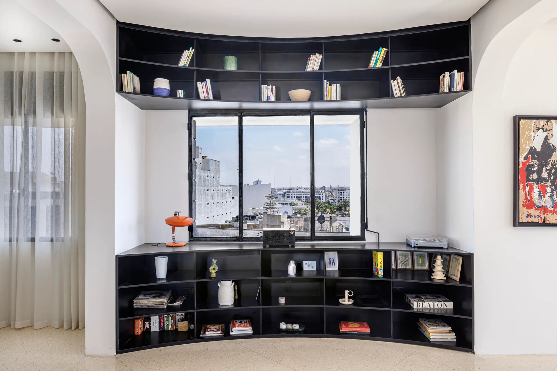 A custom curved black shelving unit built around a central window in a bright white room, displaying books and decorative objects.