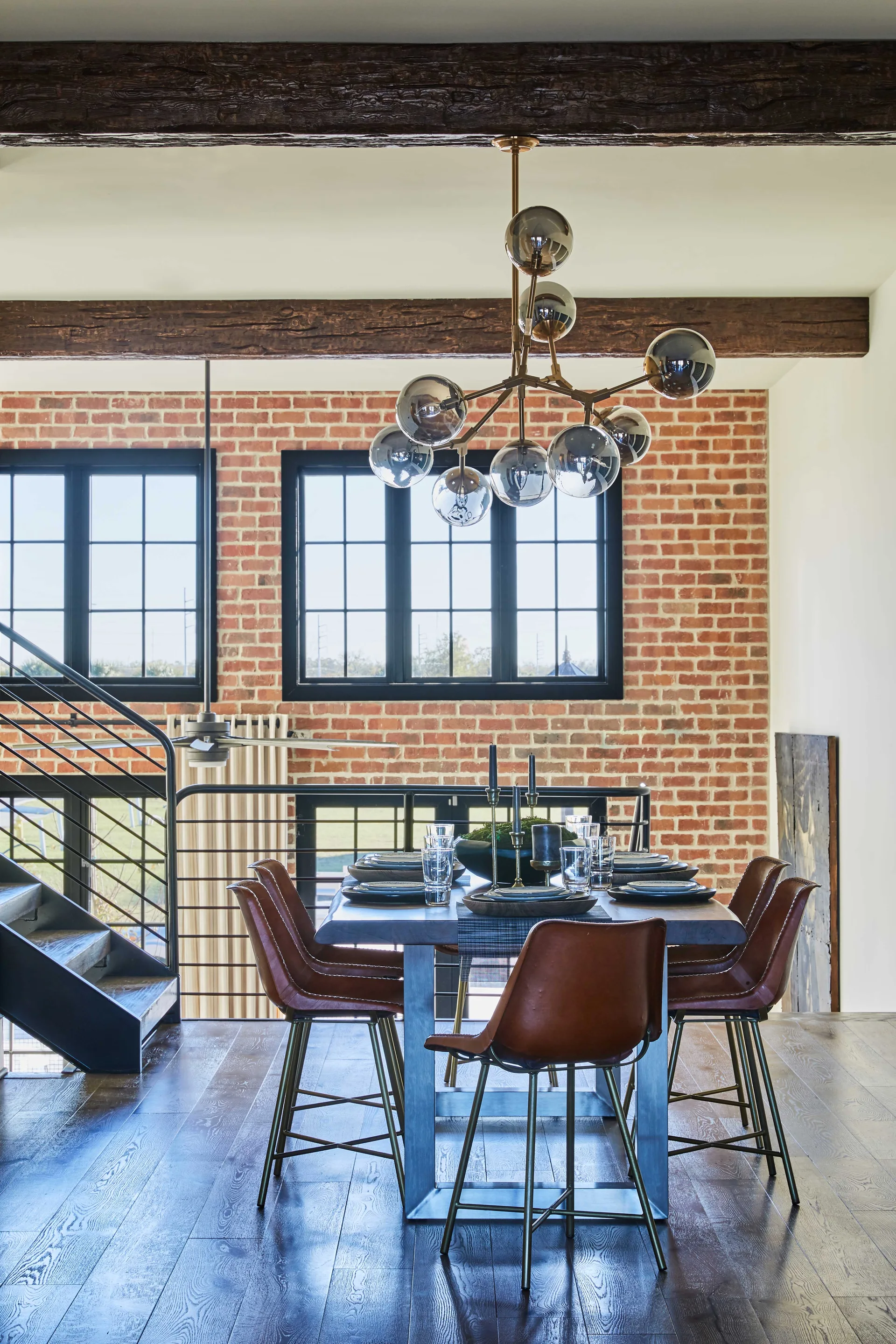 A loft dining room features a dark wood table with brown leather chairs set against an exposed red brick wall under dark ceiling beams.