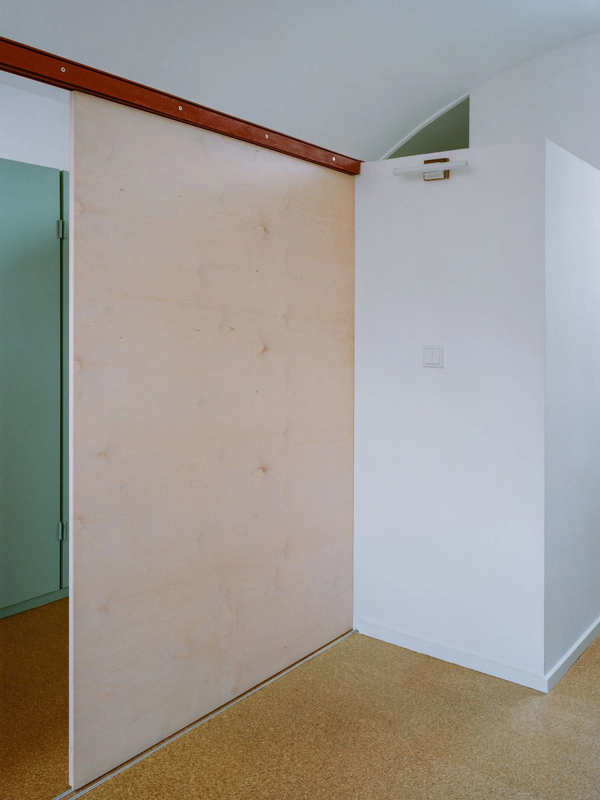 A large light-toned plywood sliding door is mounted on a rust-colored track next to a white wall and cork flooring.